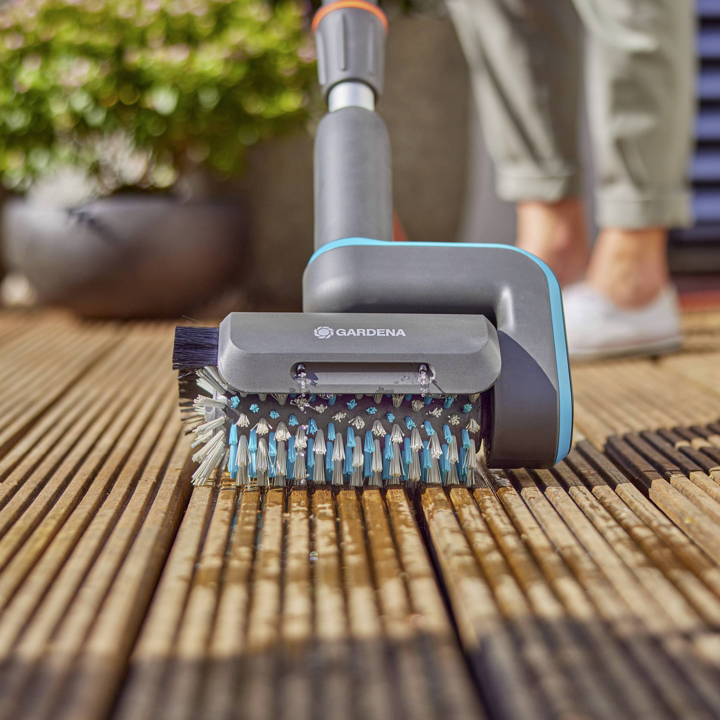 Une personne nettoie une terrasse en bois avec une brosse de nettoyage électrique de la marque Gardena. Des plantes sont visibles en arrière-plan.