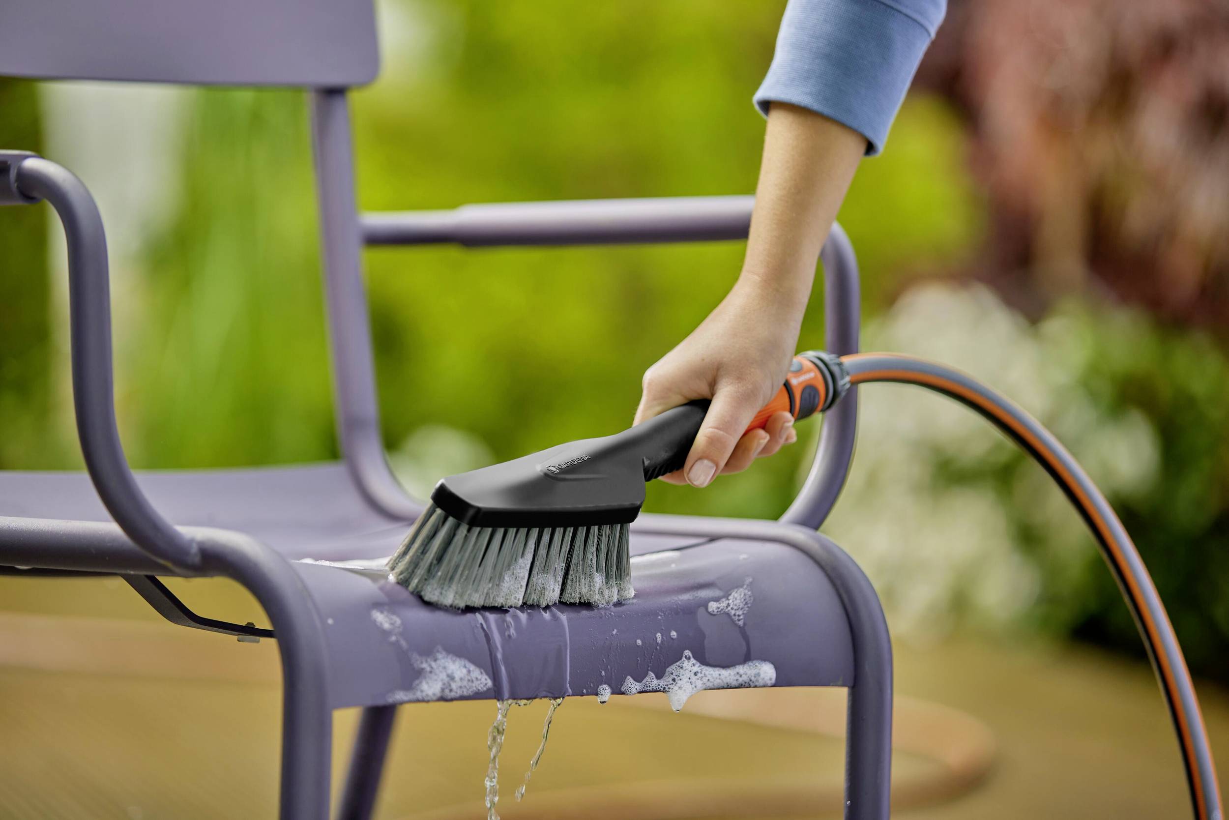 Une personne nettoie une chaise de jardin violette avec une brosse et de l'eau à l'extérieur. De la mousse est visible sur le siège.