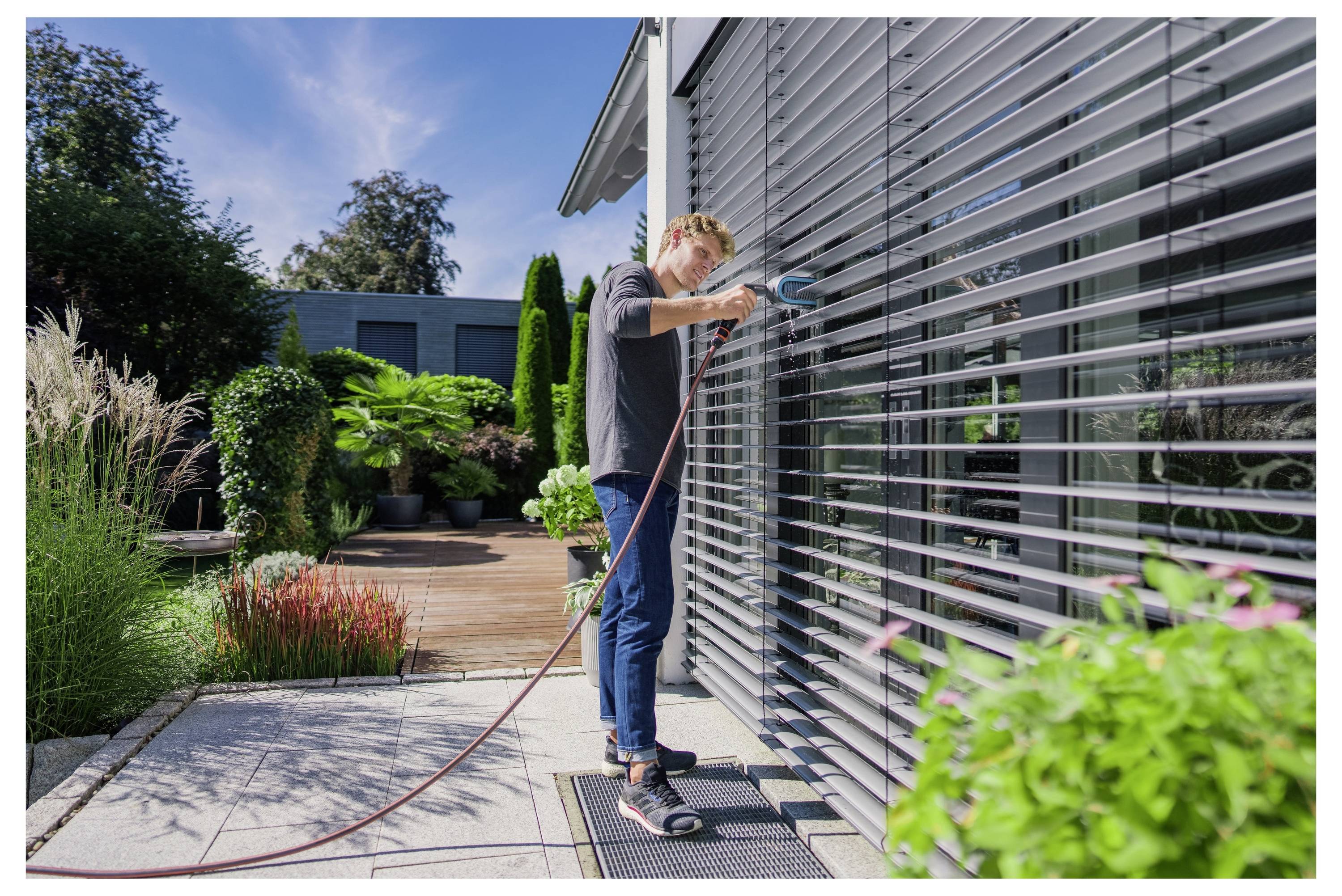Une personne nettoyant de grands stores de fenêtre avec un tuyau à l'extérieur d'une maison moderne, entourée d'un jardin et d'une terrasse.