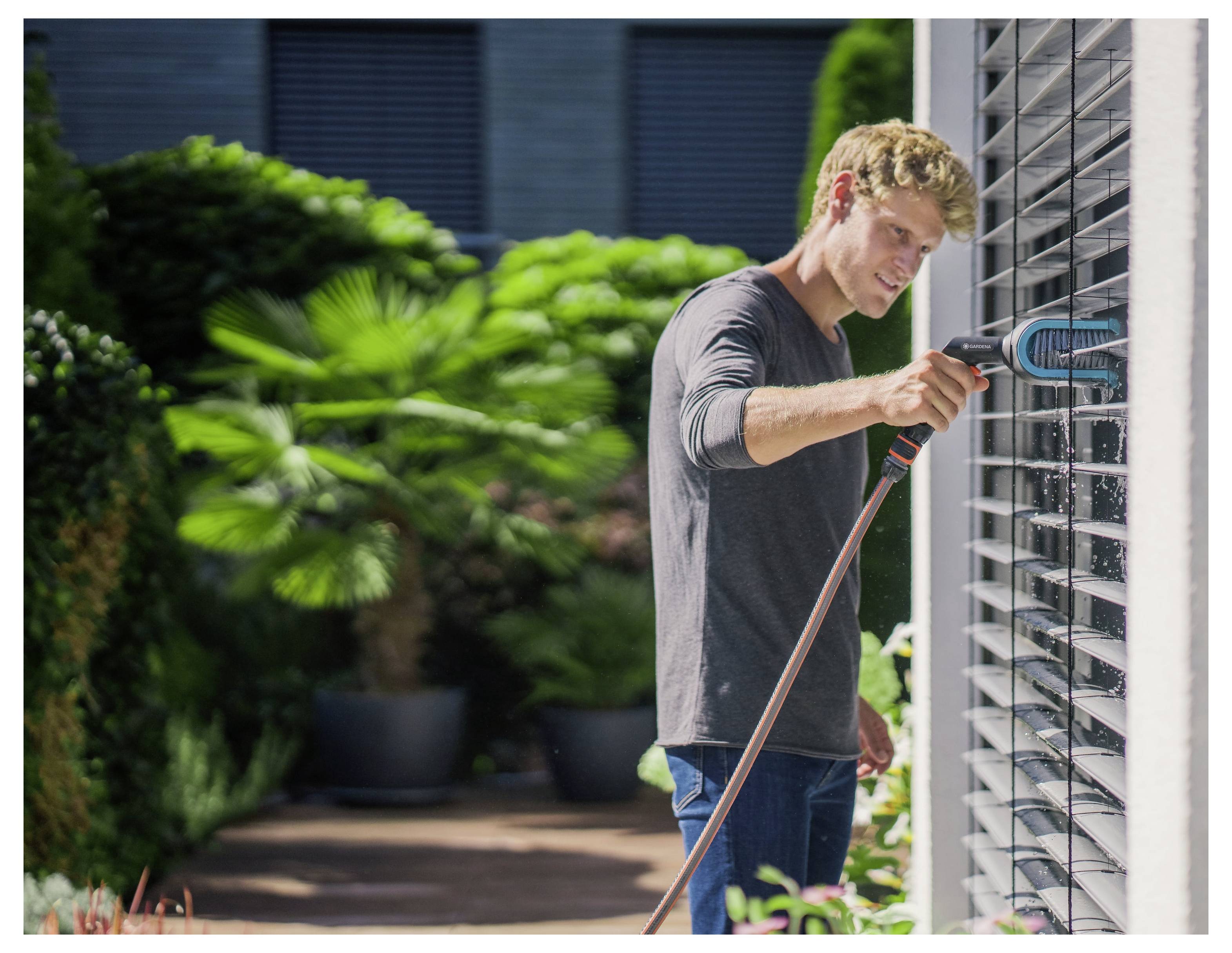 Une personne nettoie les stores extérieurs avec une brosse et un tuyau d'arrosage. Des plantes vertes luxuriantes sont visibles en arrière-plan.