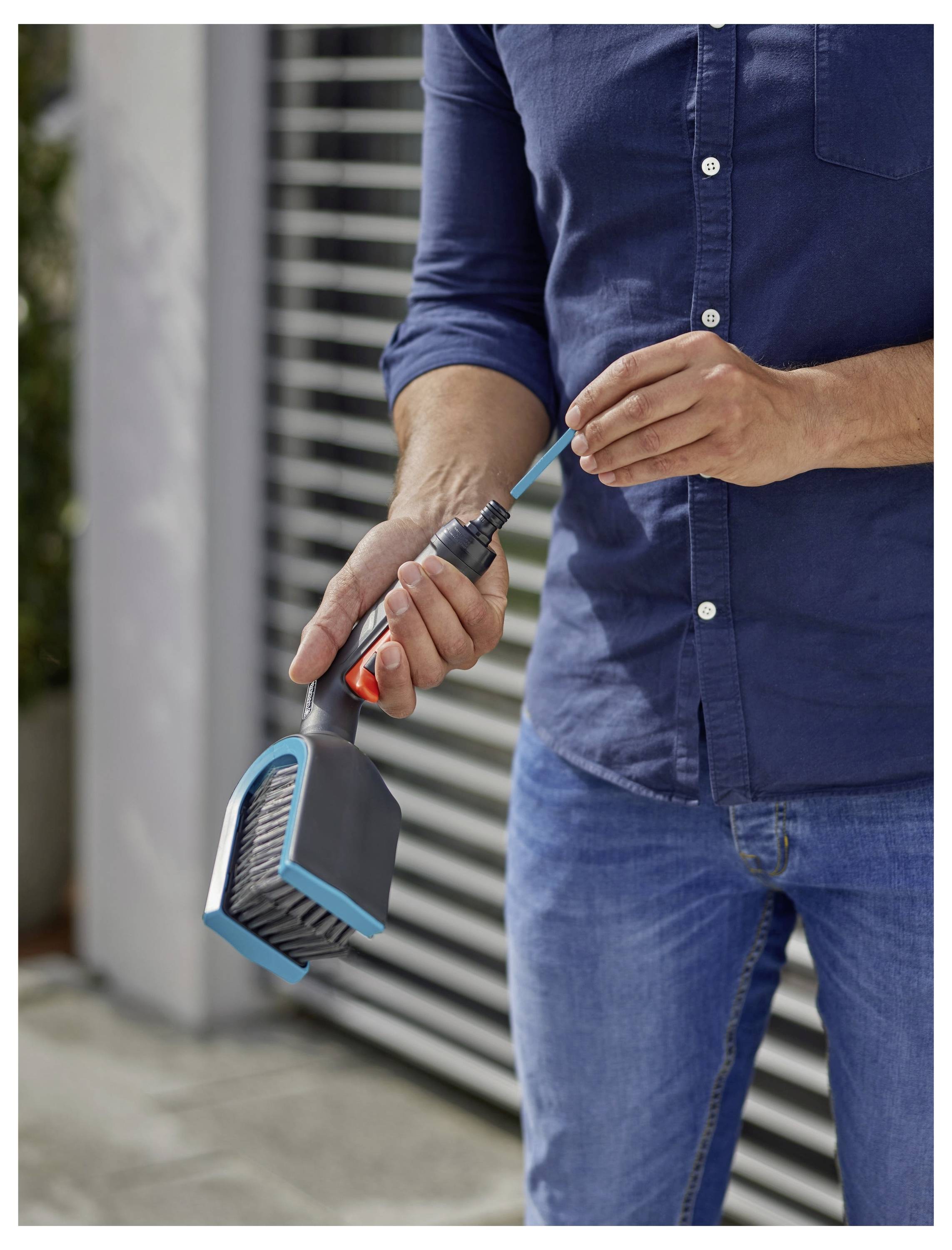 Une personne vêtue d'une chemise bleue connecte un outil de brosse à un tuyau d'arrosage pour le nettoyage, debout à l'extérieur devant un bâtiment moderne.