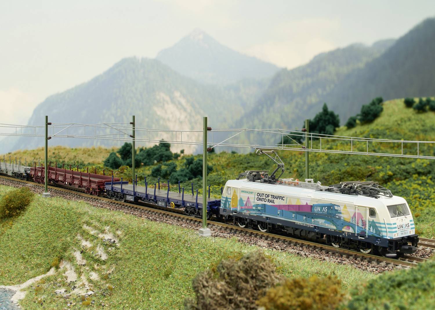Un train de marchandises traverse un paysage montagneux. Des montagnes vertes et un ciel nuageux sont visibles en arrière-plan.