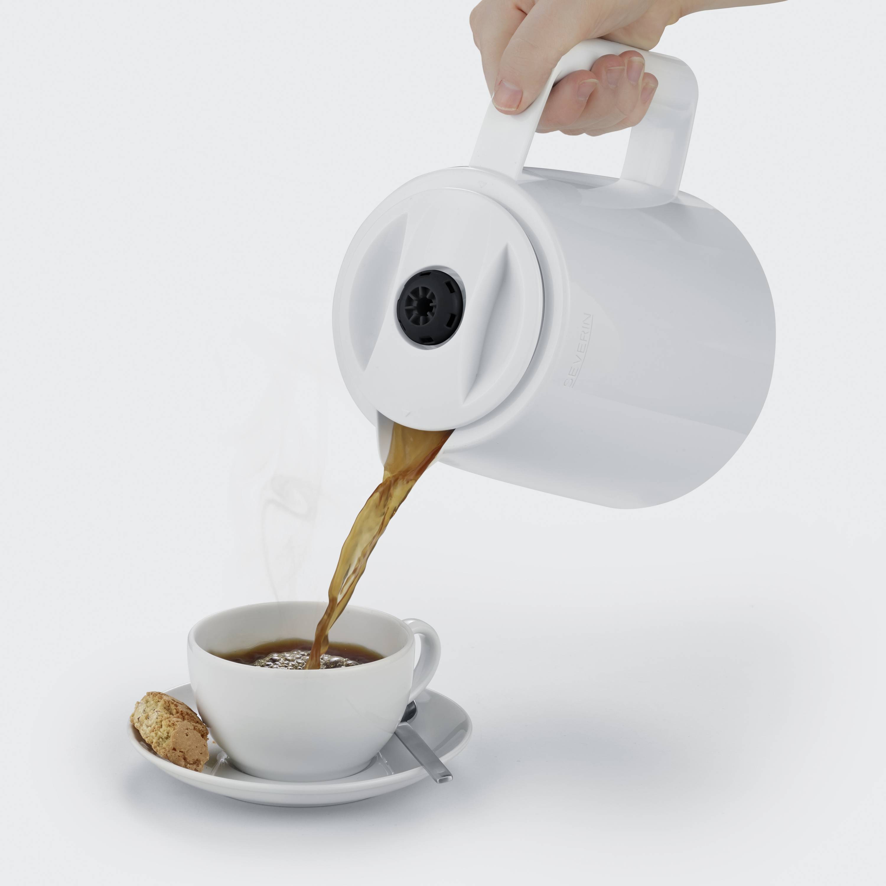 Une main verse du café d'un thermos blanc dans une tasse accompagnée d'un biscuit et d'une cuillère sur la soucoupe.