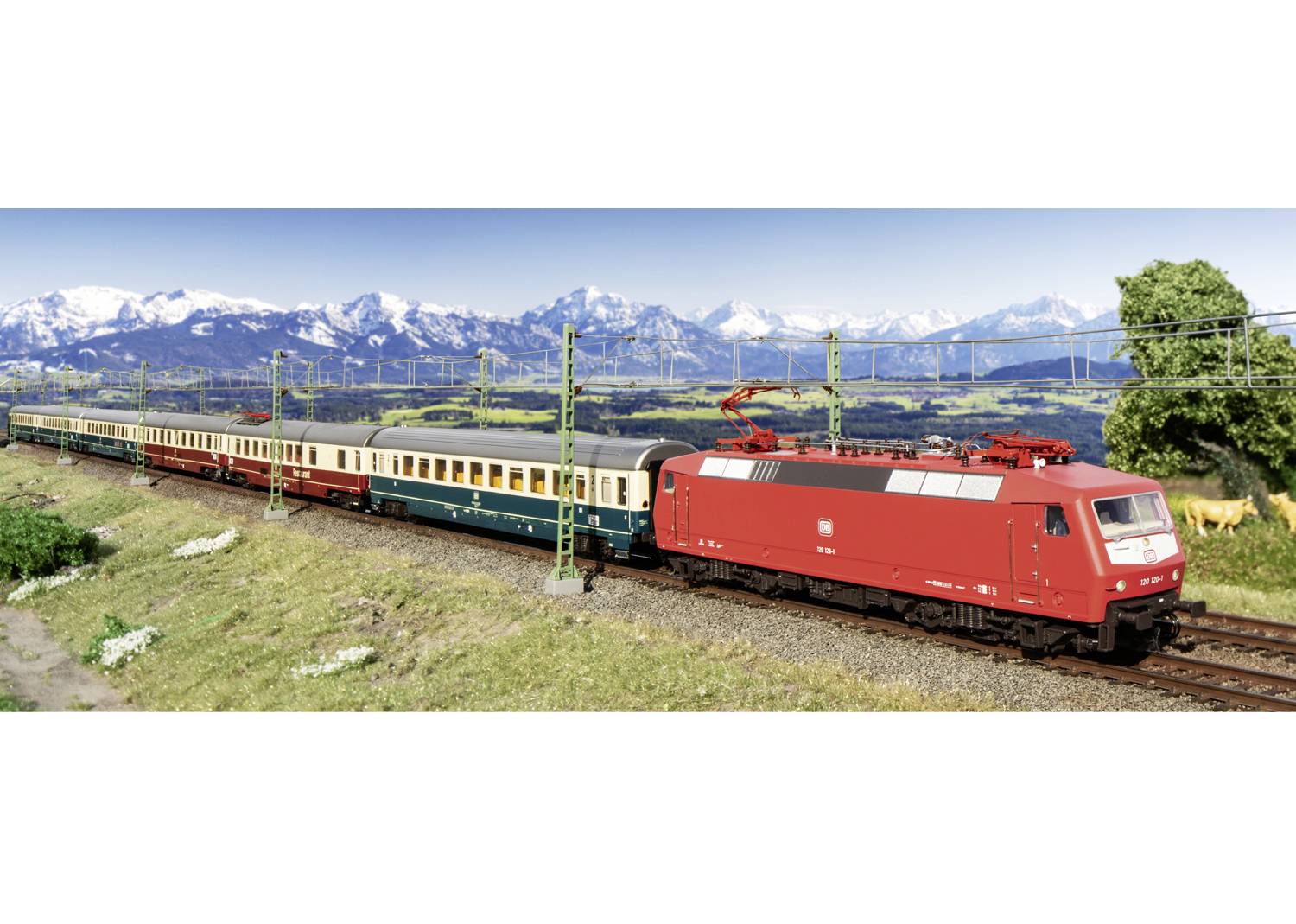 Un train rouge traverse un paysage verdoyant avec des montagnes enneigées en arrière-plan. Des lignes électriques surplombent le train.
