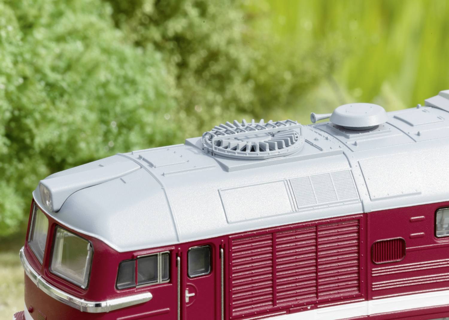 Un train miniature au corps rouge et au toit gris est placé devant un fond vert. L'accent est mis sur les détails du toit du train.