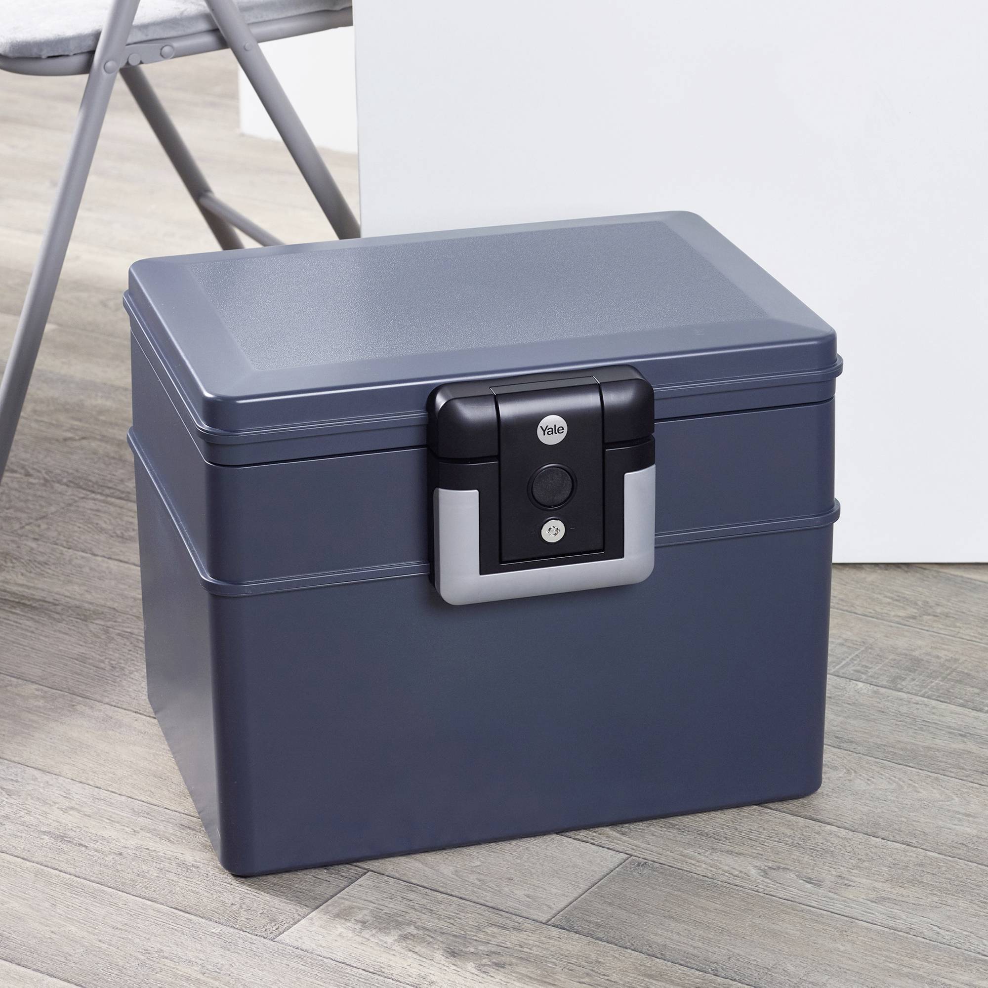 Une boîte noire, refermable, est posée sur un plancher en bois à côté d'une table blanche et d'une chaise pliante.