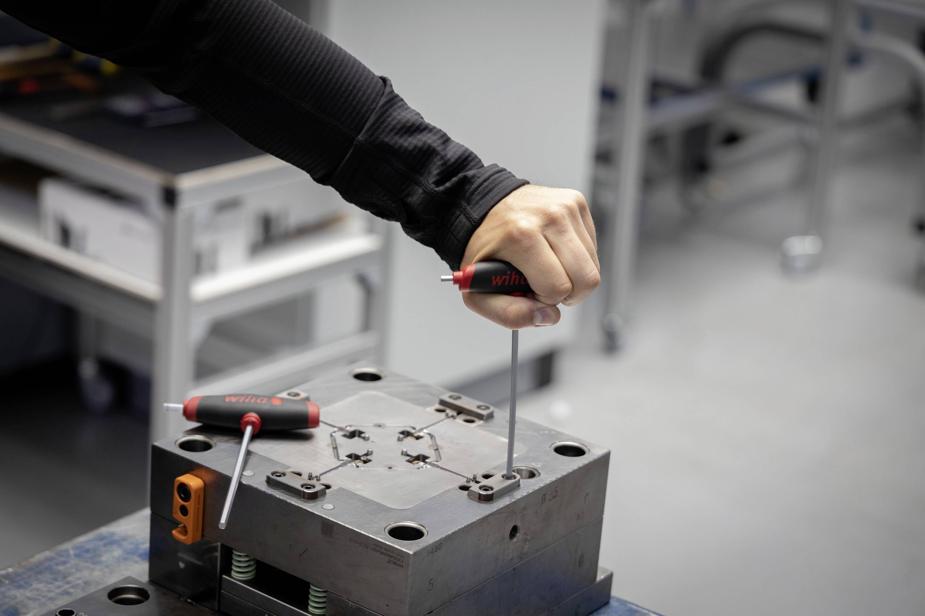 Une personne serre des vis sur une forme métallique à l'aide d'un tournevis dans un atelier. Des outils sont visibles en arrière-plan.