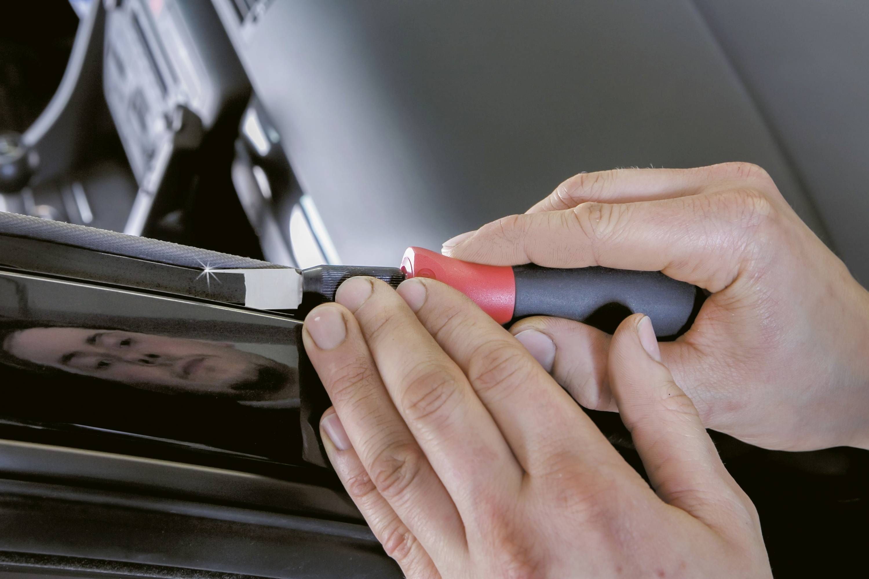 Deux mains utilisent un outil pour travailler sur le bord d'un véhicule. La scène montre un travail manuel précis sur une automobile.