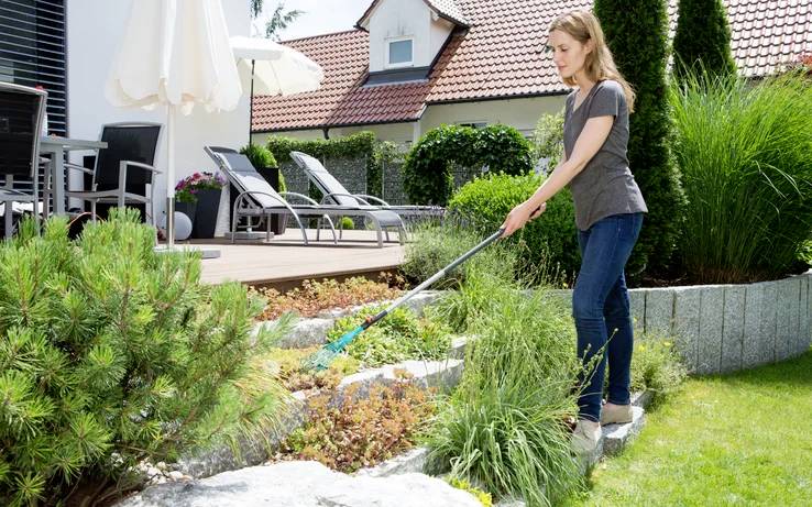 Une femme ratisse un massif de jardin dans une cour aménagée avec une terrasse, des chaises et un parasol en arrière-plan.