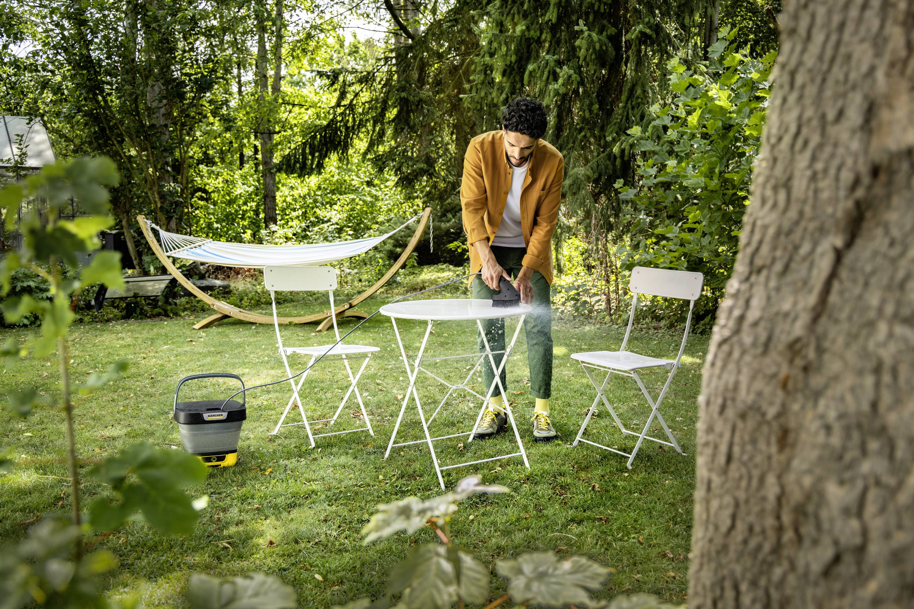 Une personne allume un barbecue sur une table dans un jardin, entourée de chaises. Une hamac est visible en arrière-plan.