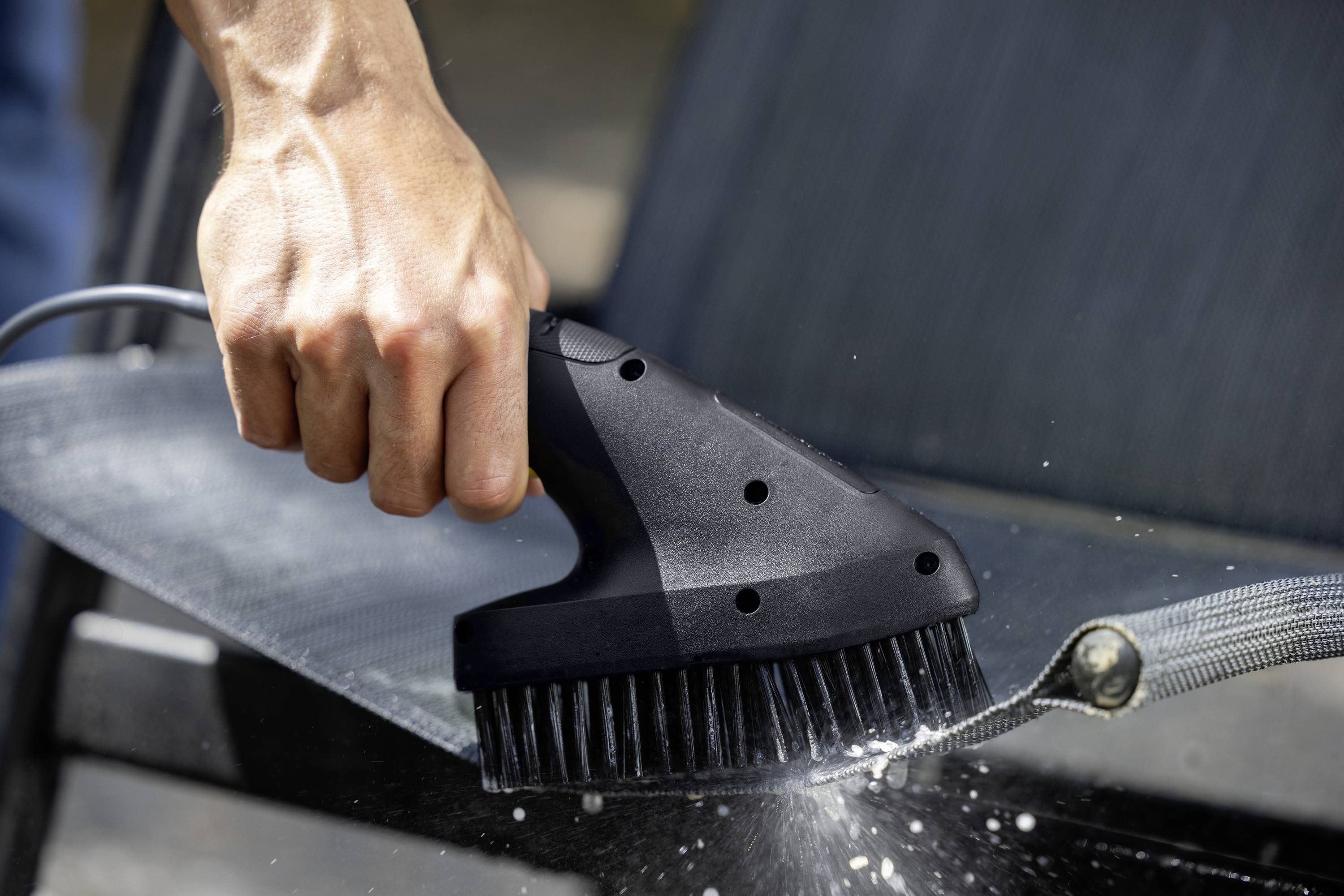 Une personne nettoie le tissu d'une chaise de jardin avec une brosse électrique. L'eau gicle de la brosse.