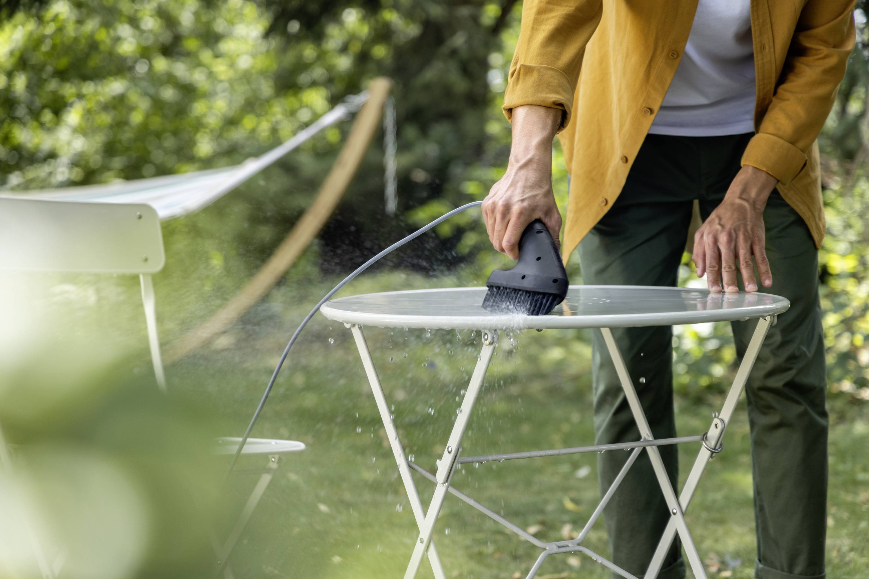 Une personne nettoie une table à l'extérieur à l'aide d'un nettoyeur vapeur à main. Un chaise blanche et de la végétation verte sont visibles en arrière-plan.