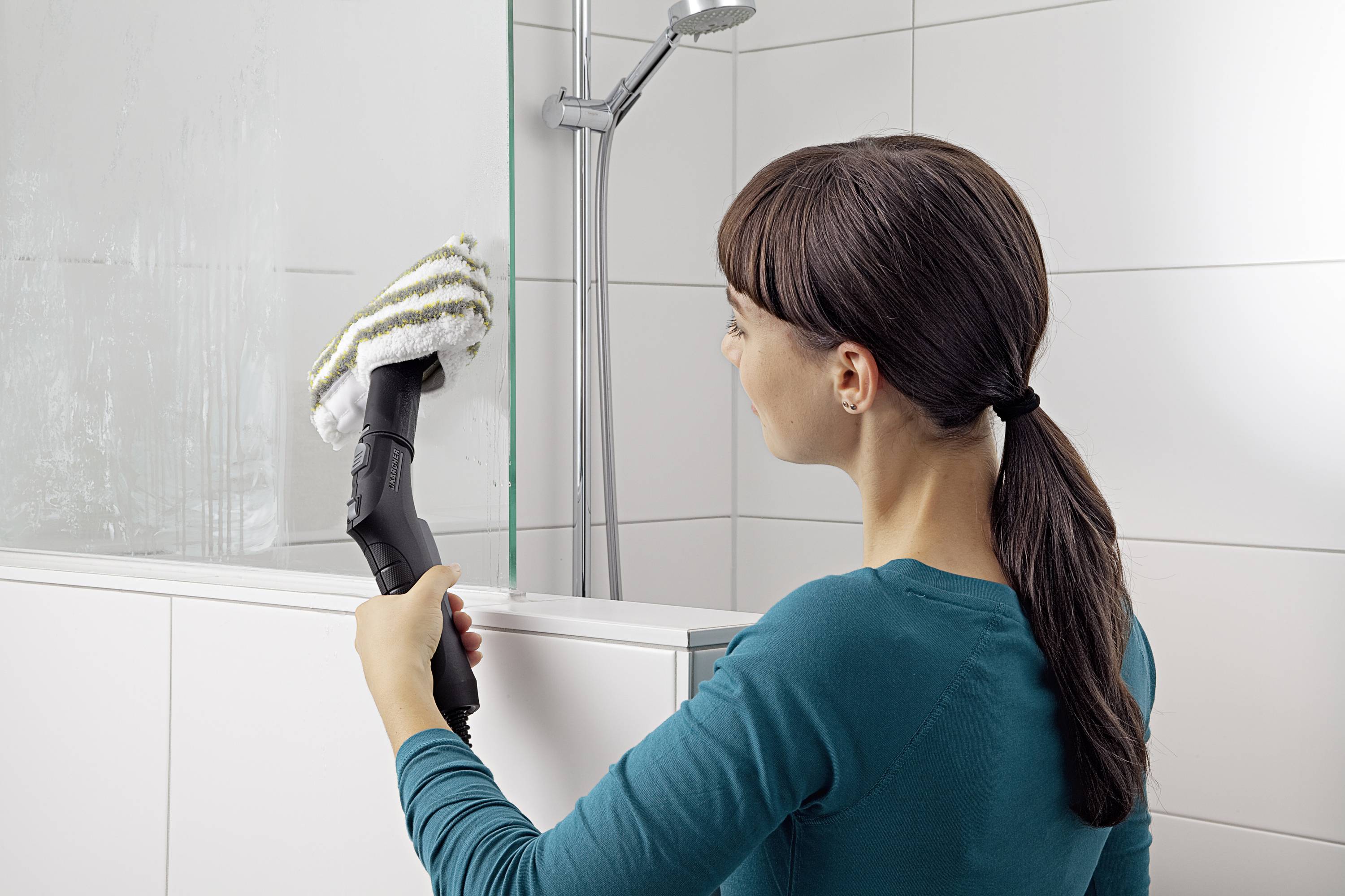 Une femme nettoie la paroi vitrée d'une cabine de douche dans une salle de bain à l'aide d'un nettoyeur vapeur. Elle porte un haut de couleur turquoise.