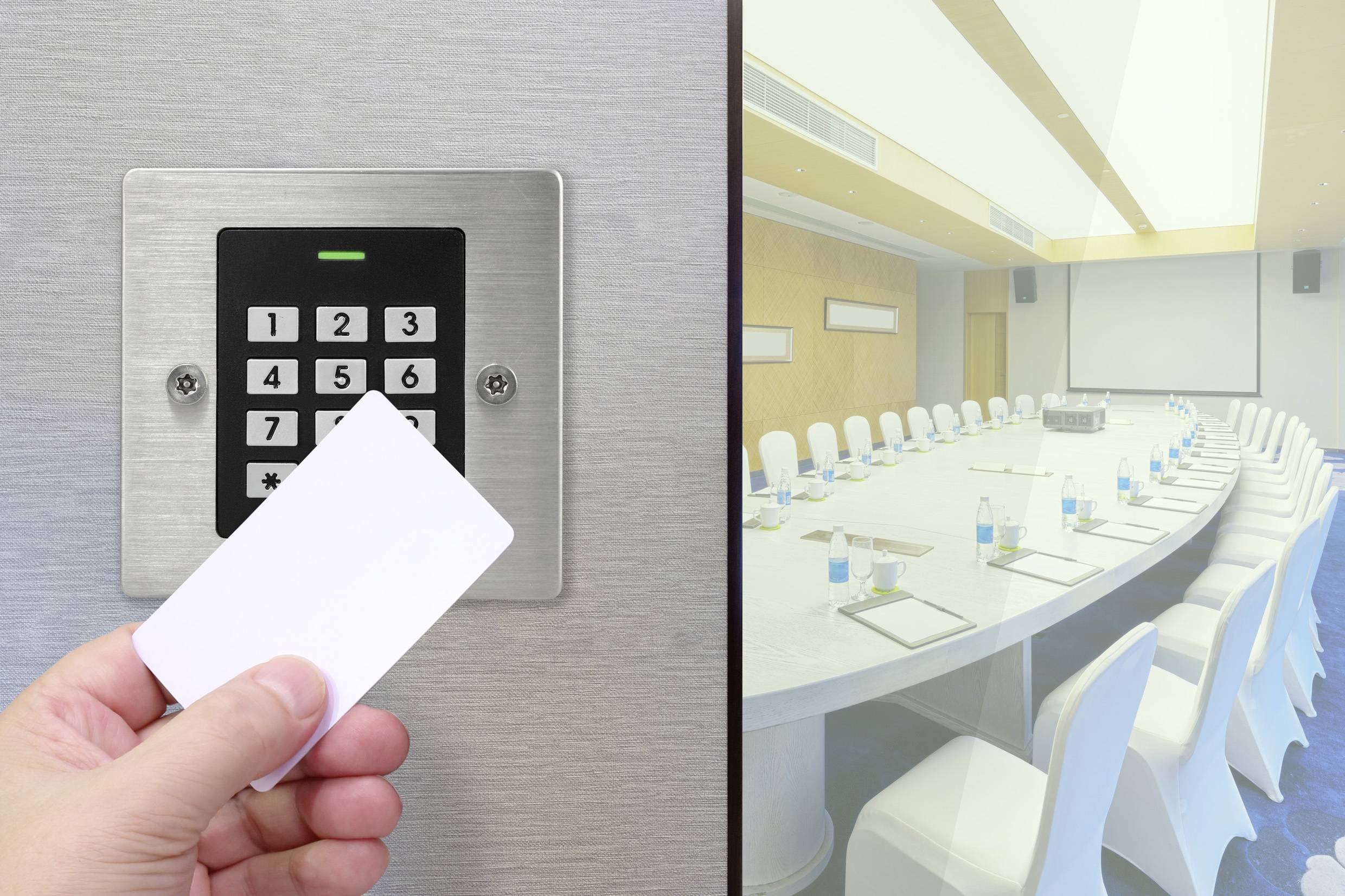 Une main tenant une carte d'accès devant un clavier de commande, à côté d'une salle de conférence moderne et vide avec une grande table ovale et des chaises blanches.