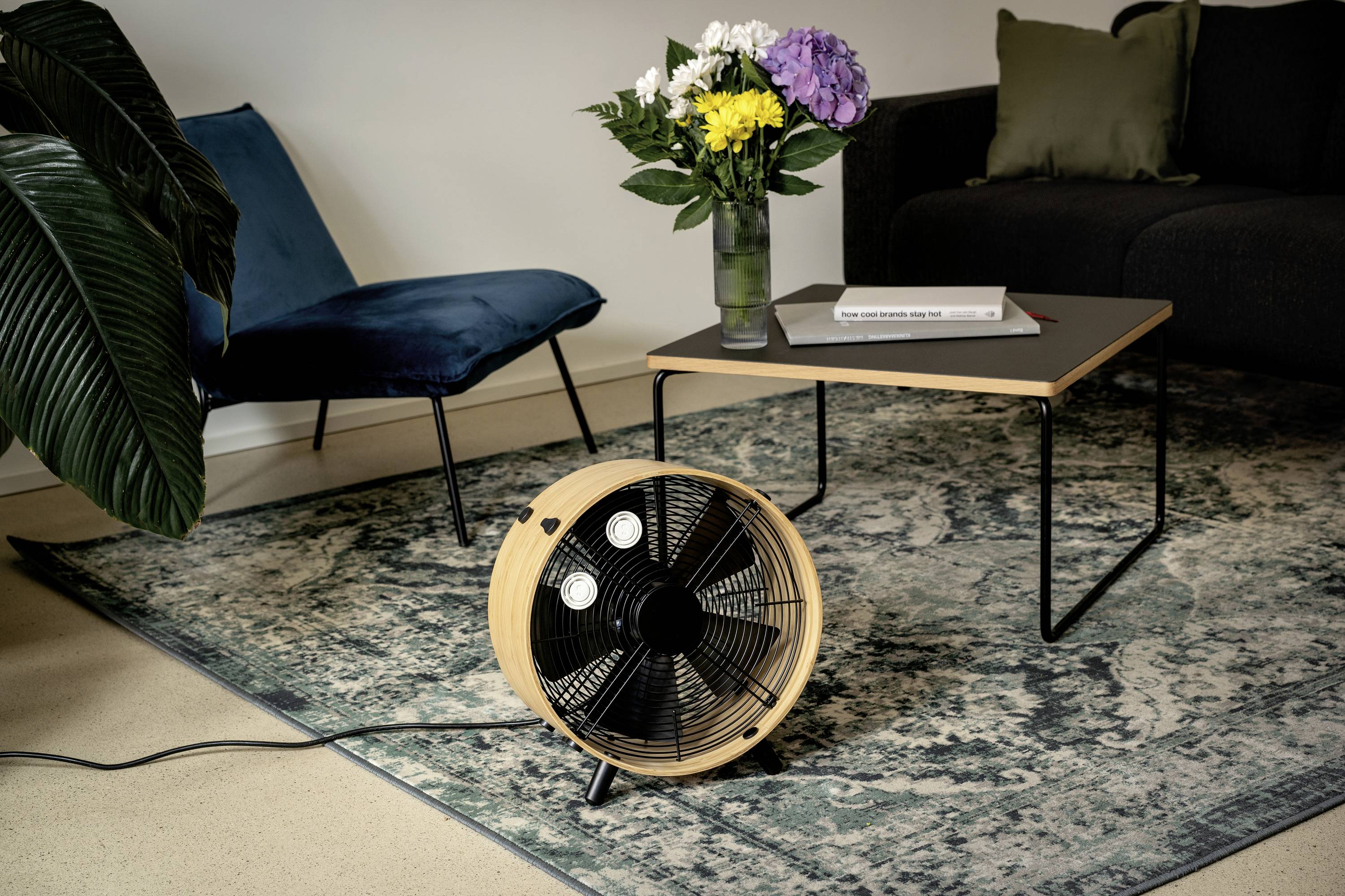 Un ventilateur sur un tapis dans un salon, à côté d'une table avec des fleurs et un livre. Un canapé et une chaise sont visibles en arrière-plan.