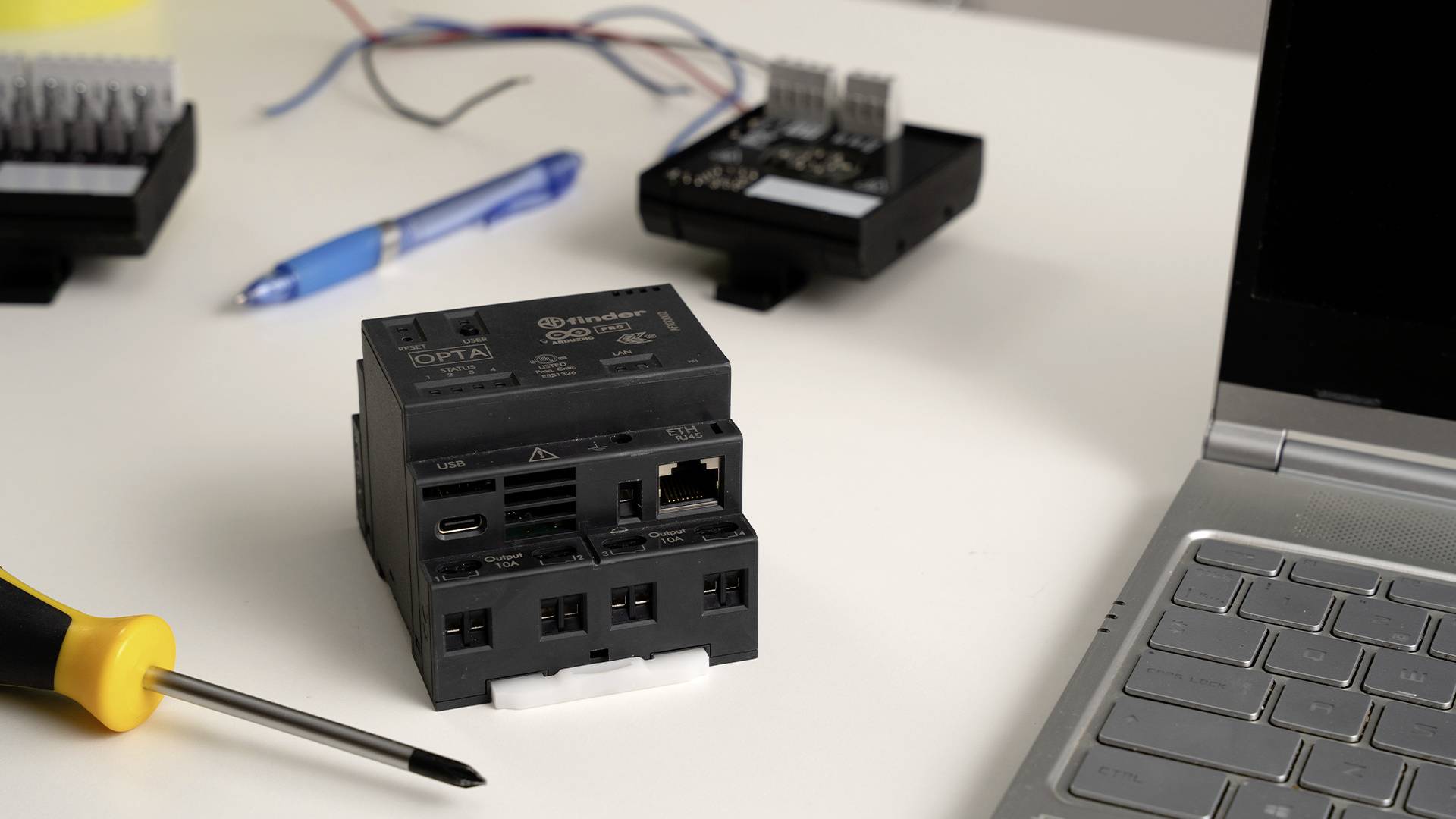 Sur une table, un appareil électrique noir est posé à côté d'un tournevis et d'un ordinateur portable. En arrière-plan, on peut voir des câbles et un stylo.