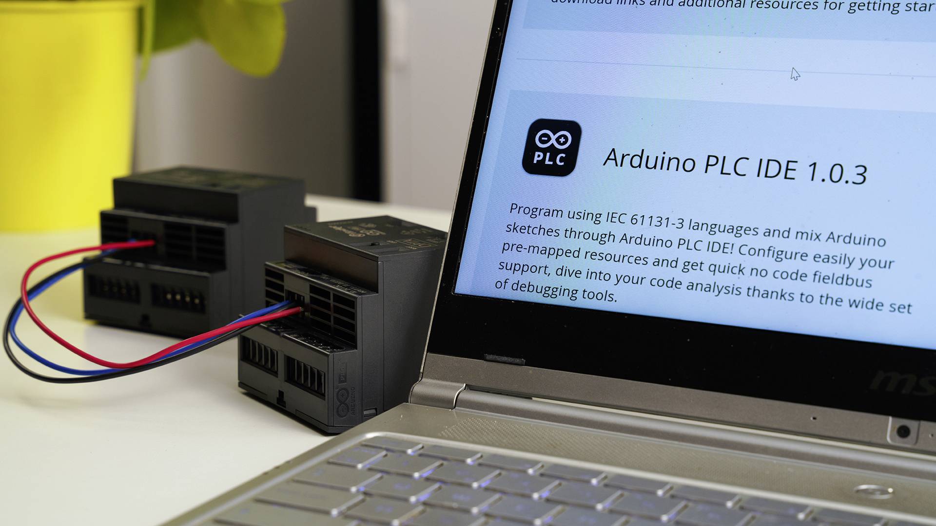 Un ordinateur portable affiche un site web avec le titre « Arduino PLC IDE 1.0.3 ». À côté se trouvent deux appareils électroniques avec des câbles.
