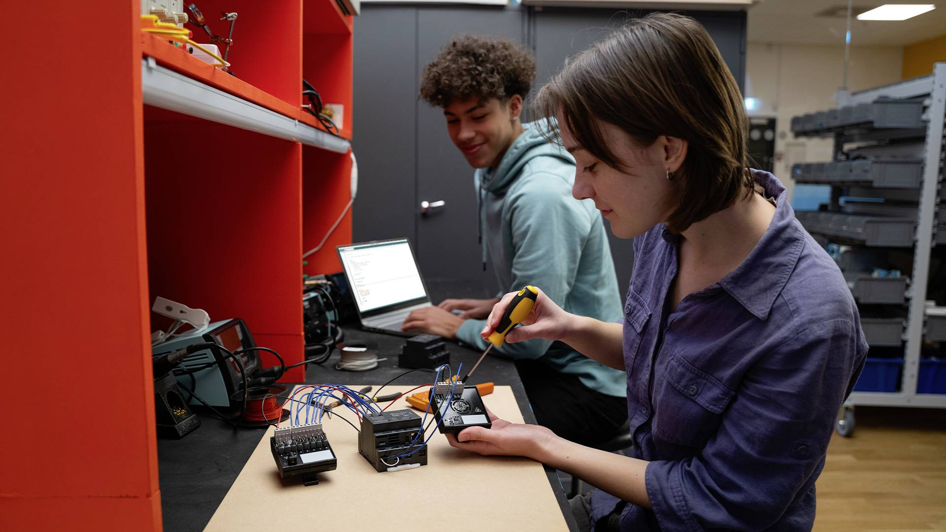 Deux personnes travaillent sur un projet électronique dans un atelier. Une personne soude, tandis que l'autre travaille sur un ordinateur portable.