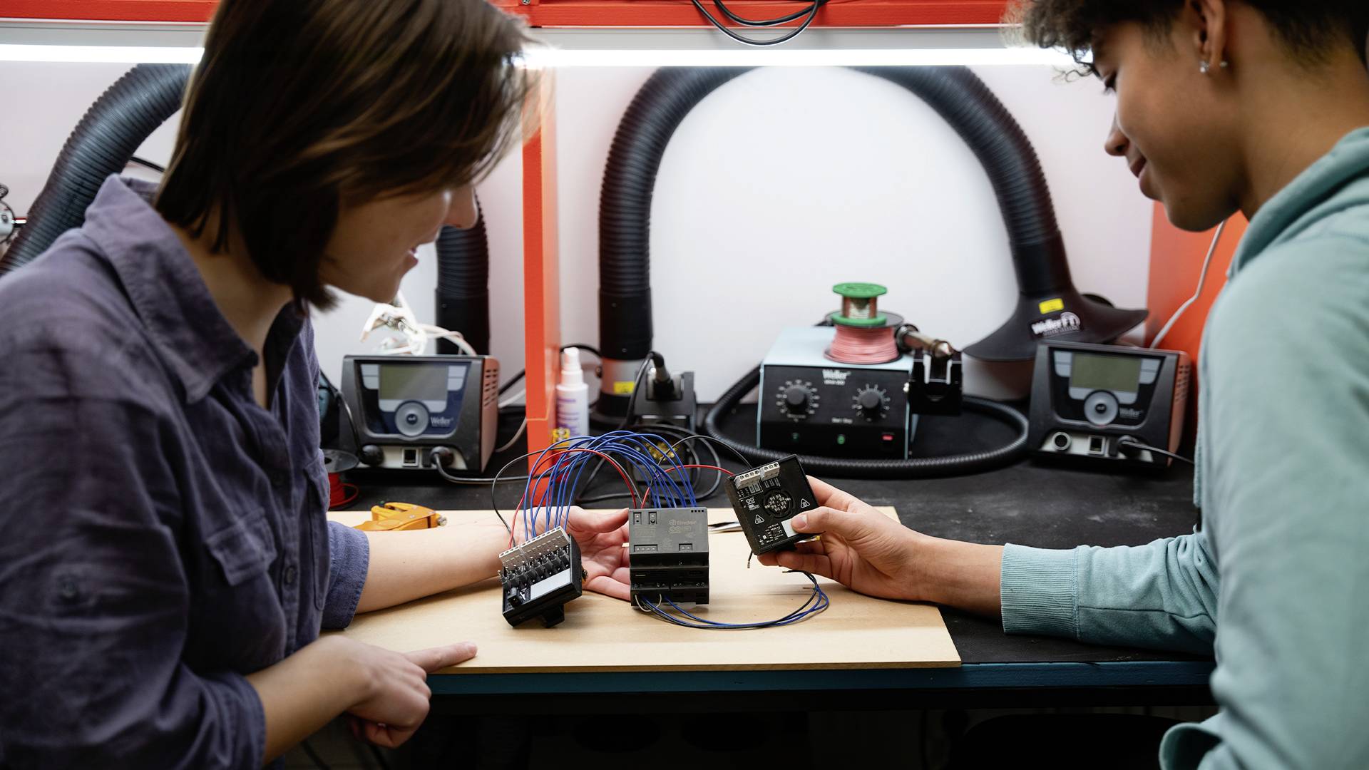 Deux personnes travaillent sur un projet de circuit électronique à une table, entourées de fers à souder et d'outils.
