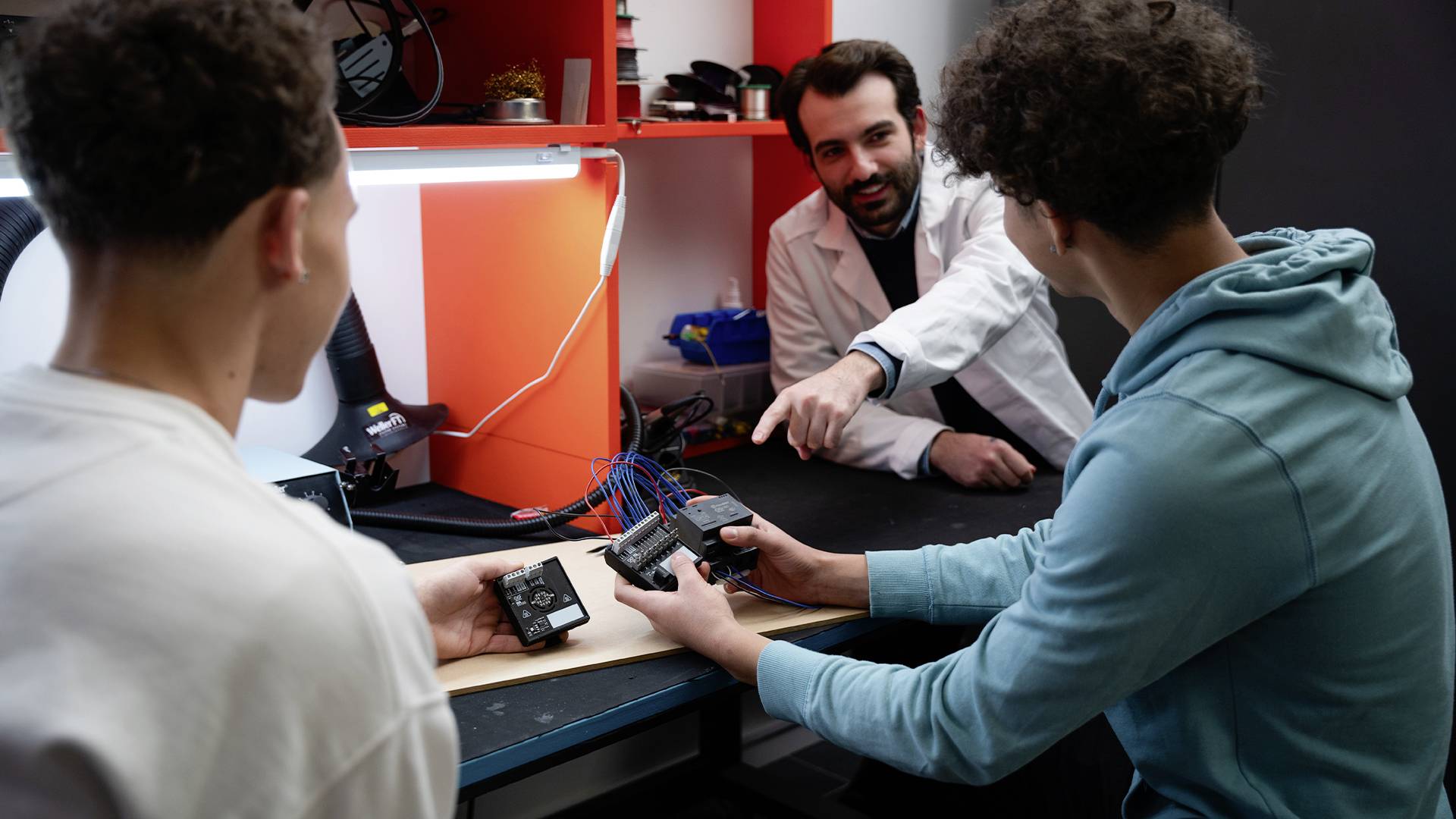Trois personnes dans un laboratoire. Deux jeunes personnes tiennent des appareils électroniques, tandis qu'un homme en blouse blanche explique.