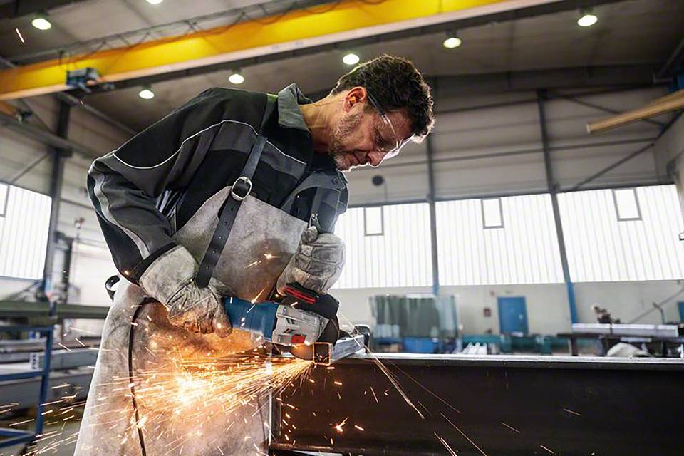 Un homme dans un atelier porte des équipements de protection et utilise un outil de meulage électrique alors que des étincelles volent.