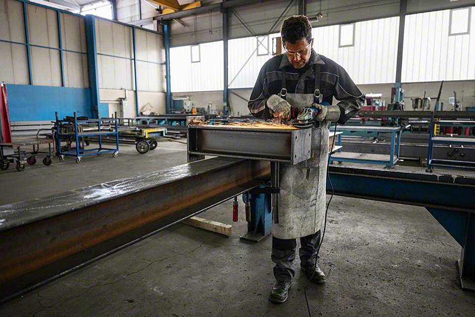Un homme travaille dans un atelier, utilisant un outil de meulage sur un grand support métallique. Des étincelles volent tout autour.