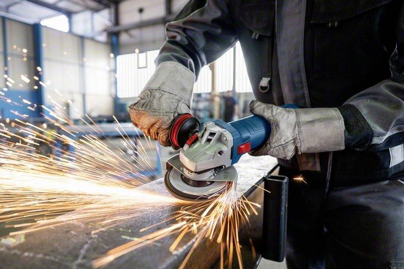 Une personne en tenue de protection meule du métal avec une meuleuse d'angle, des étincelles volent. La scène se déroule dans un atelier.
