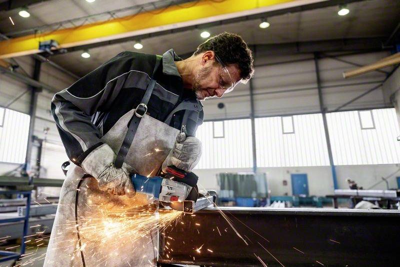Un ouvrier en tenue de protection meule du métal avec une meuleuse d'angle. Des étincelles volent tandis qu'il travaille dans un environnement industriel.