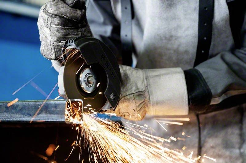Une personne coupe du métal avec une scie circulaire, des étincelles volent. Des vêtements de protection sont portés.