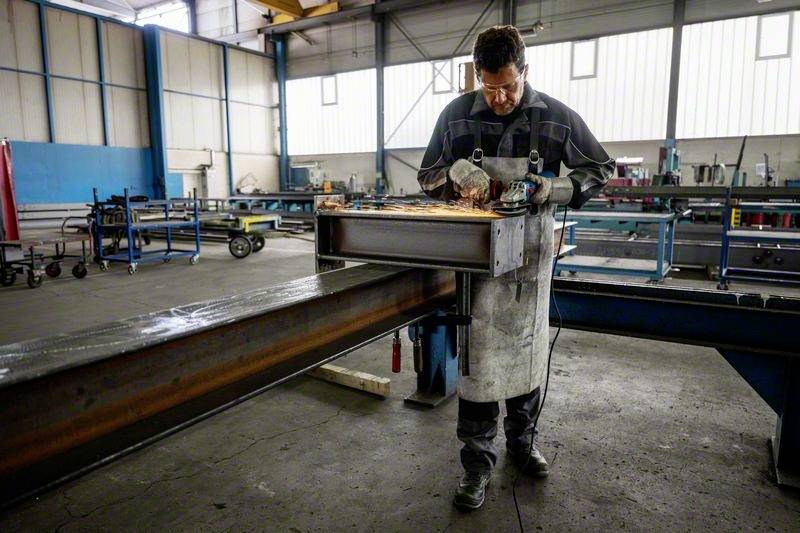 Un ouvrier meule du métal dans un hall d'usine. Il porte des lunettes de protection et un tablier. Des étincelles volent pendant le meulage. Des machines sont visibles en arrière-plan.