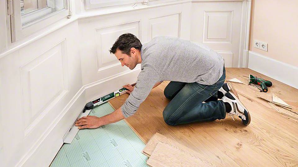 Un homme est agenouillé dans une pièce et fixe une plinthe à l'aide d'un outil. Le sol est composé de bois et de matériau d'isolation.