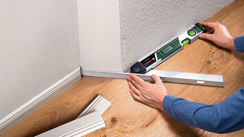 Une personne mesure la distance entre le mur et le plinthes à l'aide d'une règle et d'un niveau à bulle sur un sol en bois.