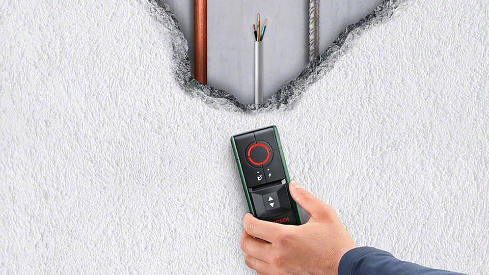 Une personne tient un appareil de mesure Bosch contre un mur dont la surface est percée, révélant des câbles et des tuyaux à l'intérieur.
