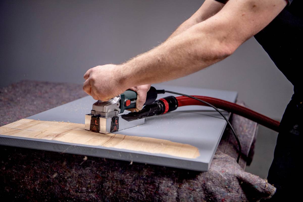 Une personne travaille une planche de bois avec un outil électrique. Des copeaux de bois sont visibles tandis que l'outil crée une surface lisse.
