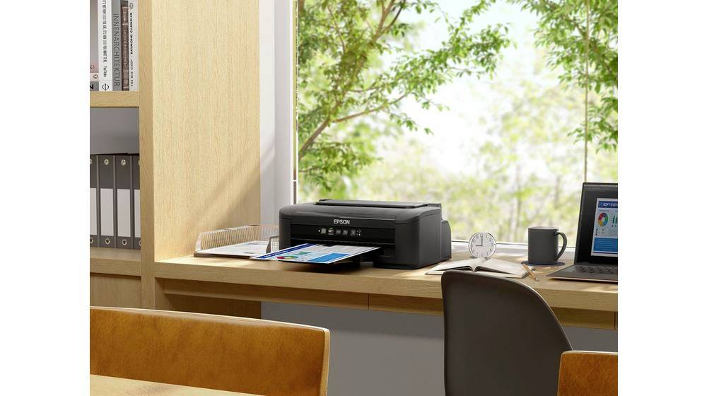 Une imprimante sur un bureau imprime un document coloré. En arrière-plan, on peut voir un ordinateur portable et une vue sur des arbres verdoyants.
