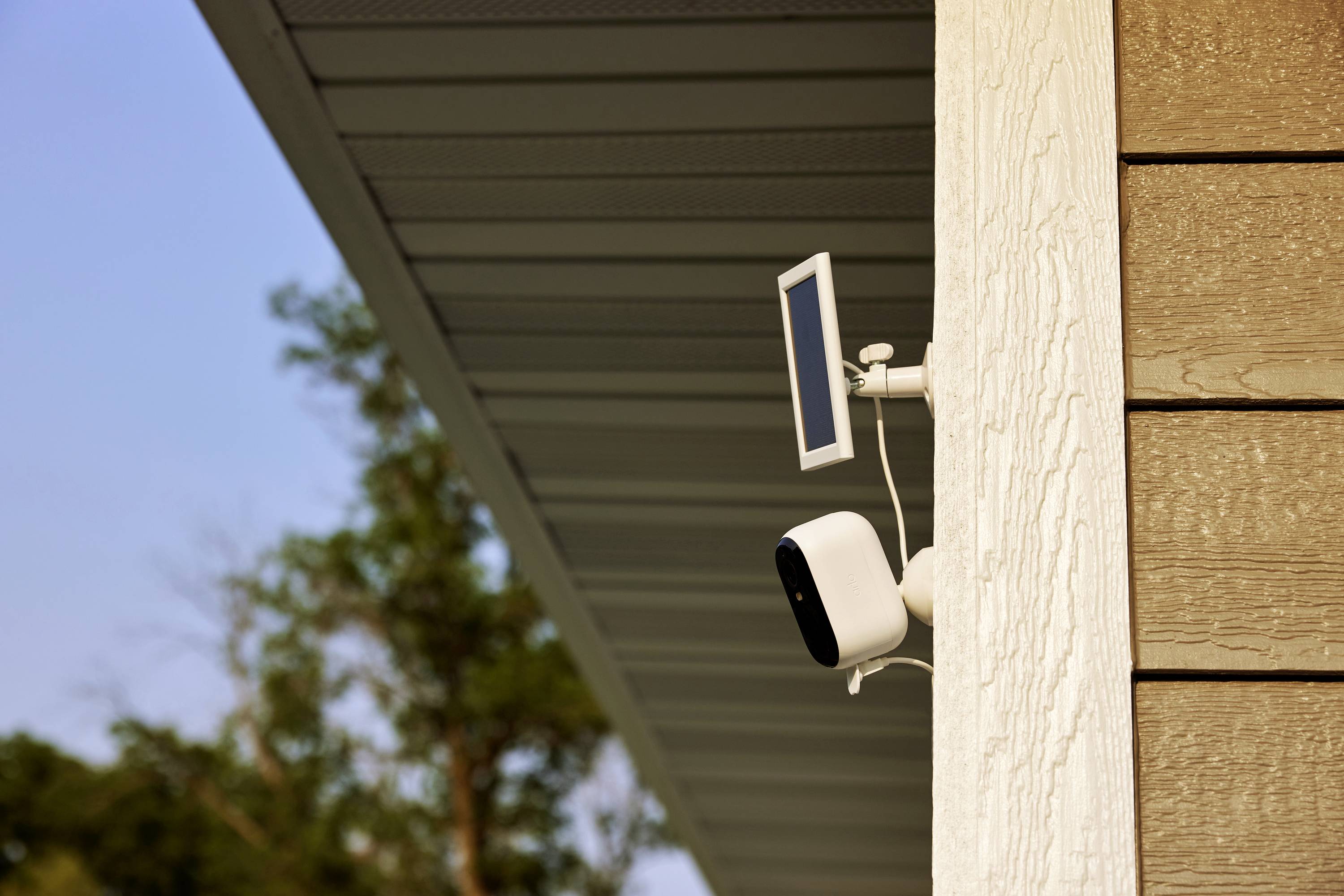 Une caméra de surveillance est fixée sur le côté extérieur d'un bâtiment, avec un petit panneau solaire juste au-dessus. Des arbres sont visibles en arrière-plan.