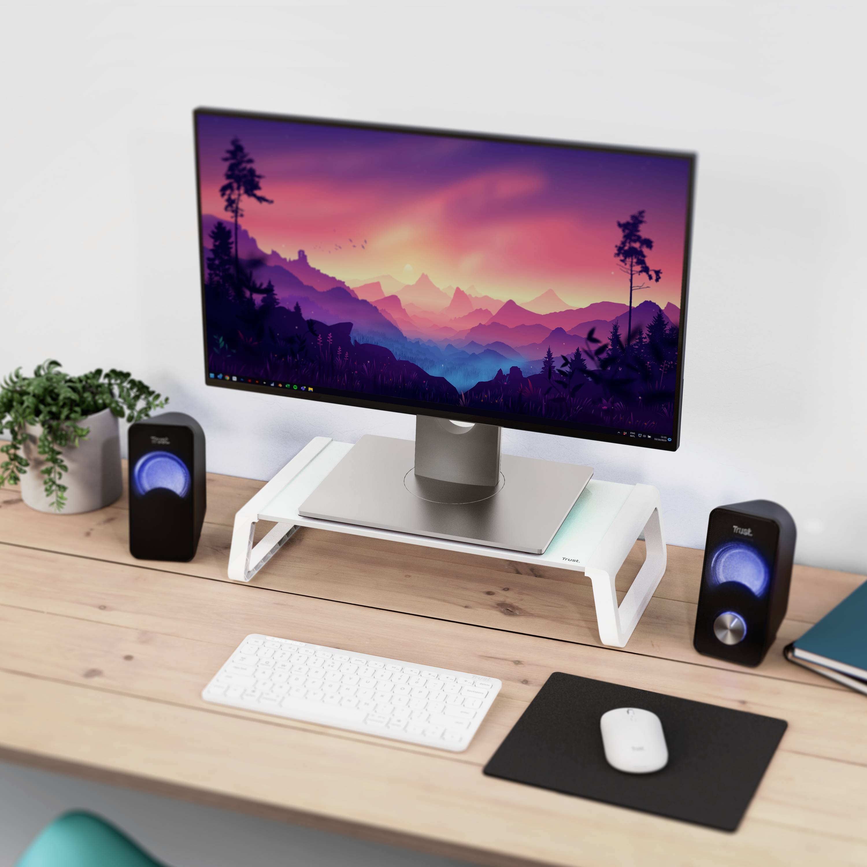 Un bureau avec un écran d'ordinateur affichant un paysage, des enceintes, un clavier, une souris et une plante en pot.