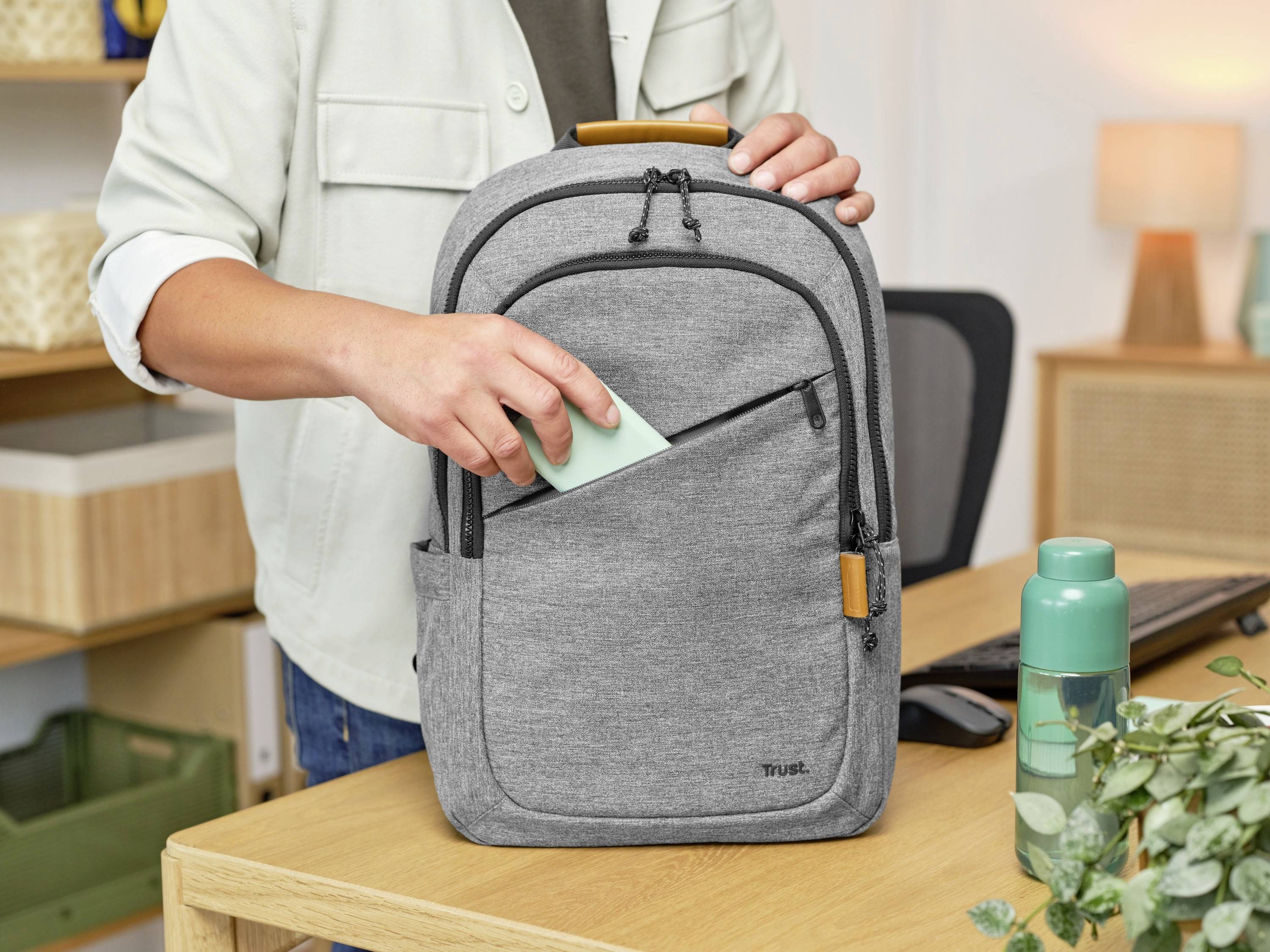 Une personne glisse une note verte dans la poche avant d'un sac à dos gris, qui est posé sur un bureau à côté d'une bouteille d'eau.