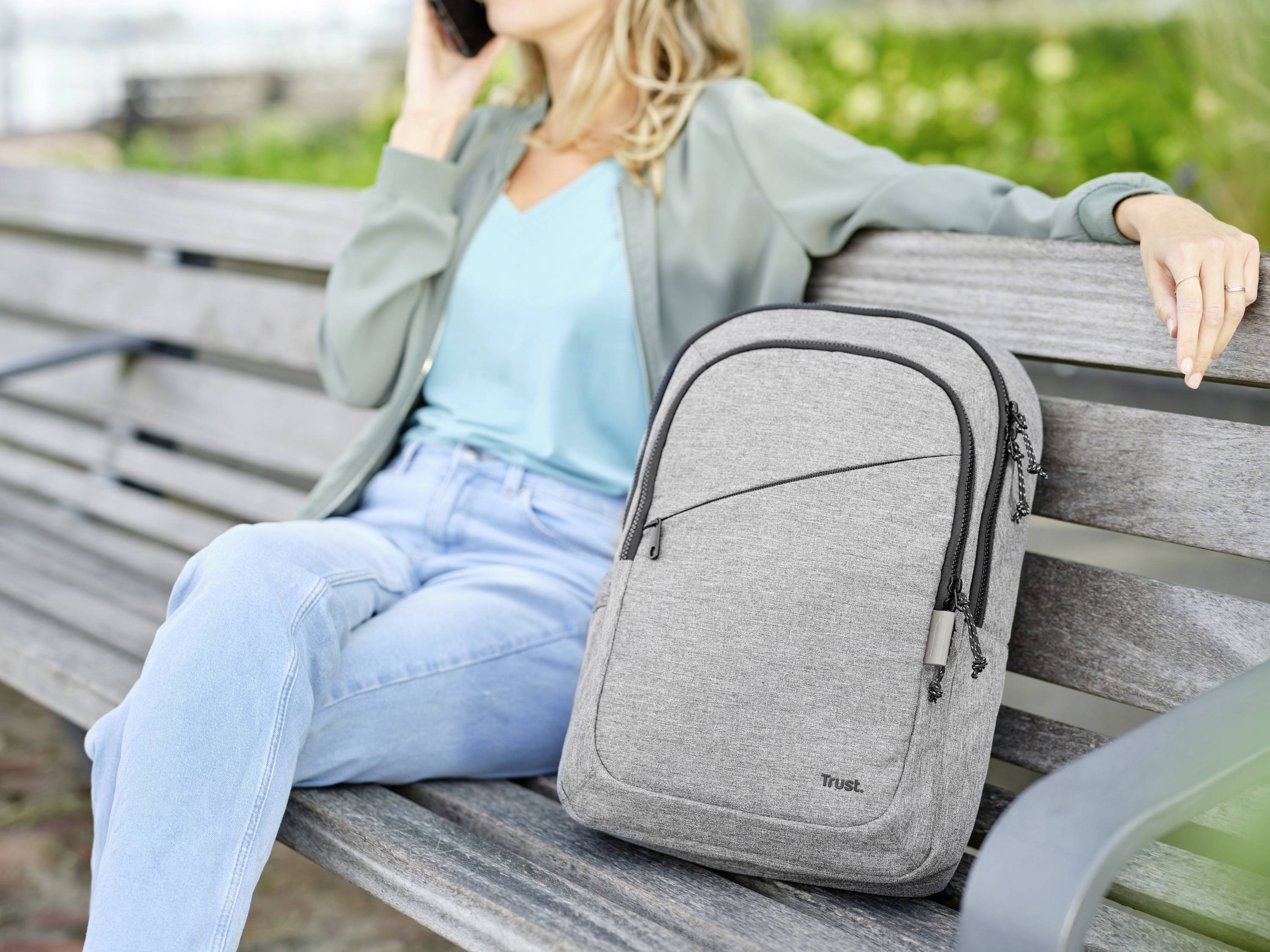 Une femme est assise souriante sur un banc de parc et téléphone, tandis qu'un sac à dos gris est posé à côté d'elle.