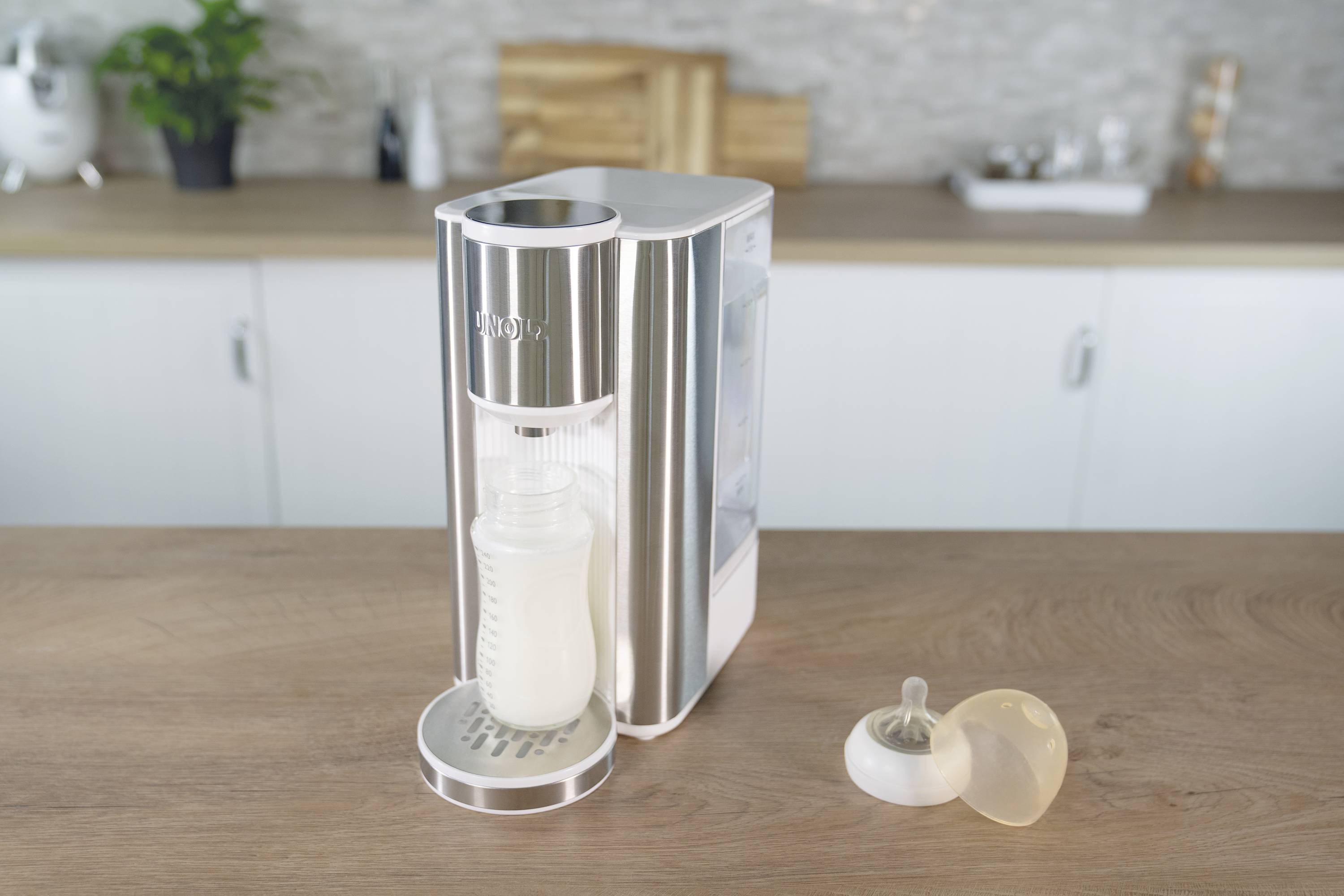 Une machine à préparer les biberons posée sur un plan de travail de cuisine, avec un récipient de lait rempli à côté. En arrière-plan, on aperçoit l'aménagement de la cuisine.