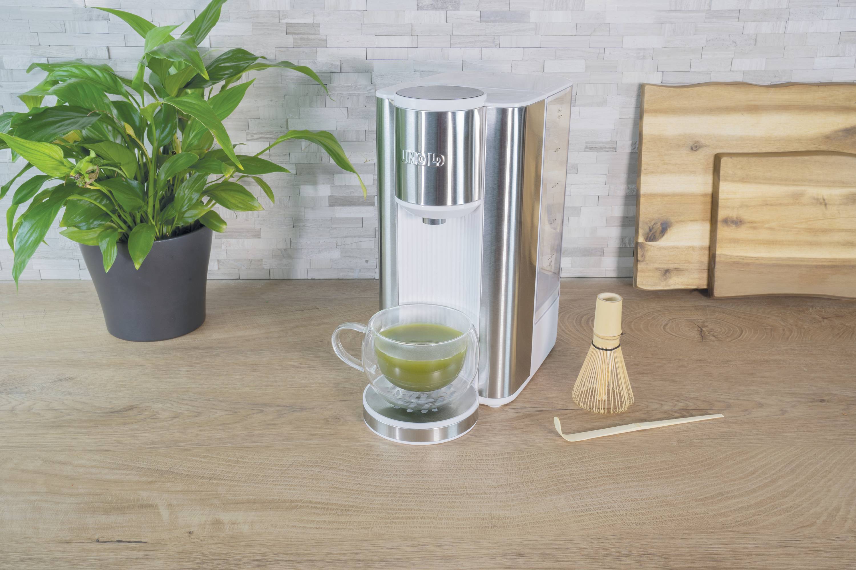 Machine à thé argentée avec une tasse de thé vert, à côté d'un fouet en bambou et d'une cuillère en bois sur une table en bois. À gauche, un pot avec une plante est posé.