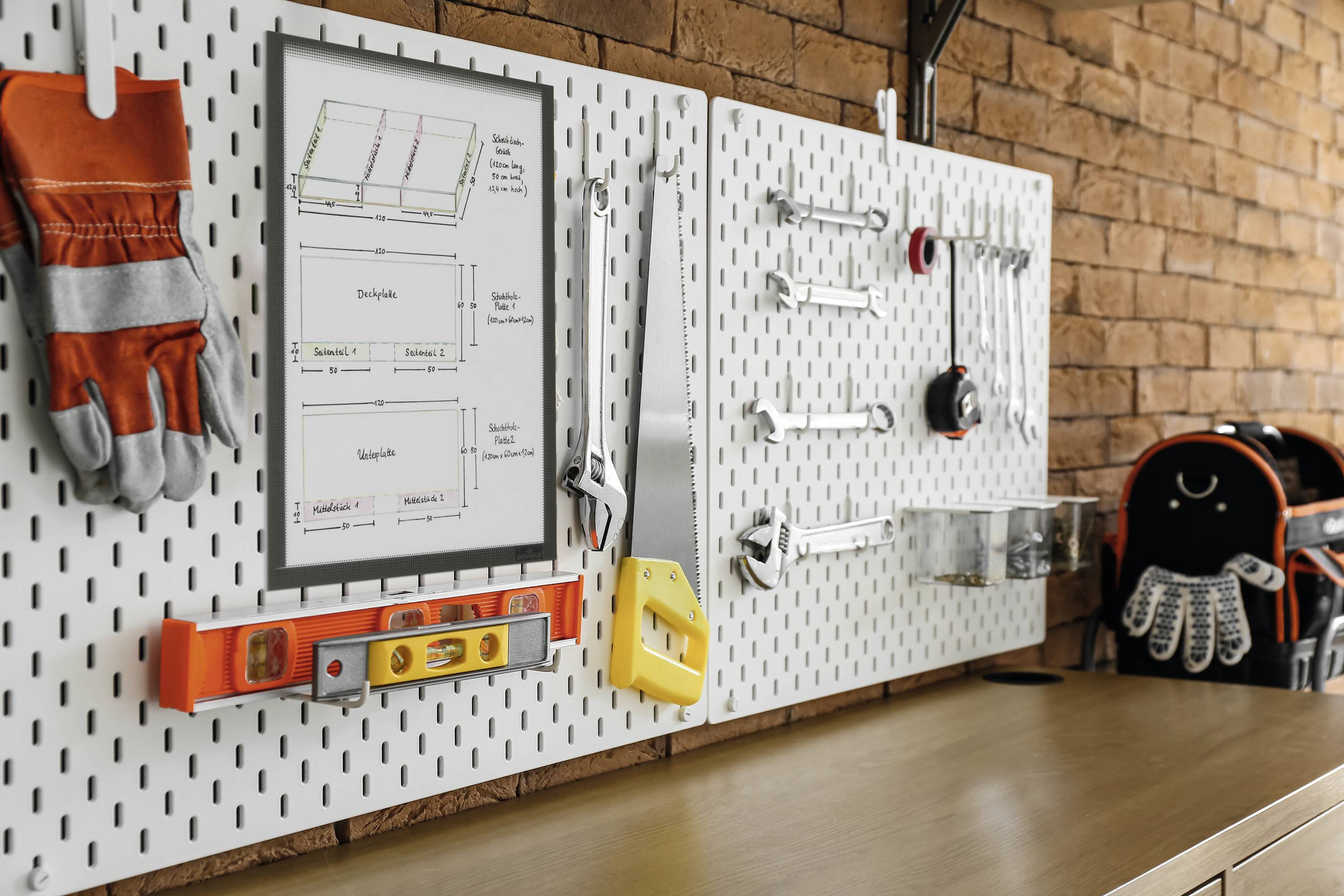 Mur d'outils avec des gants, un niveau à bulle, des clés à molette et un plan de construction, fond en briques rouges, sac à dos orange et noir à droite.