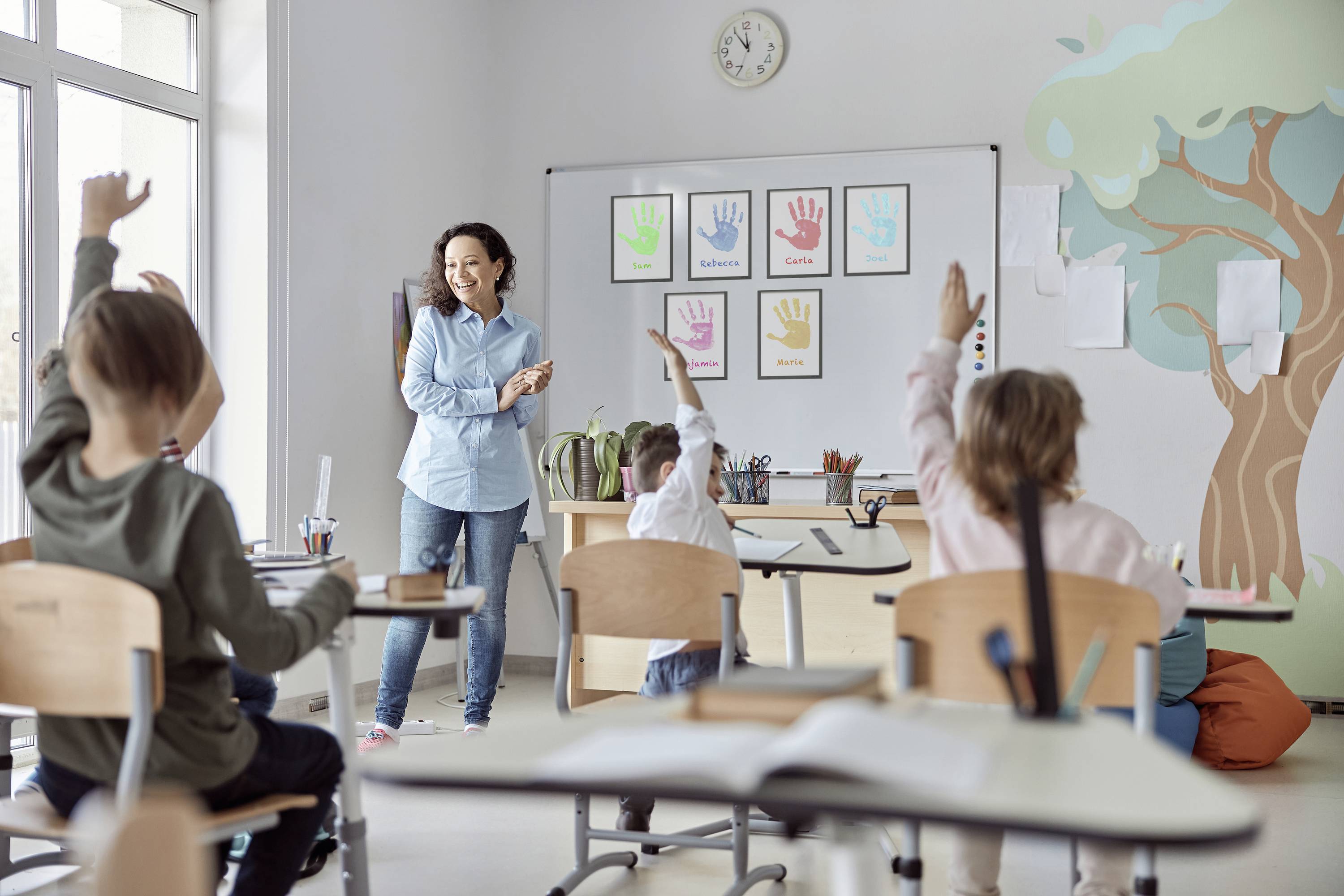 Une enseignante se tient devant une classe d'élèves, qui lèvent leurs mains. Au tableau, des empreintes de mains colorées sont affichées.