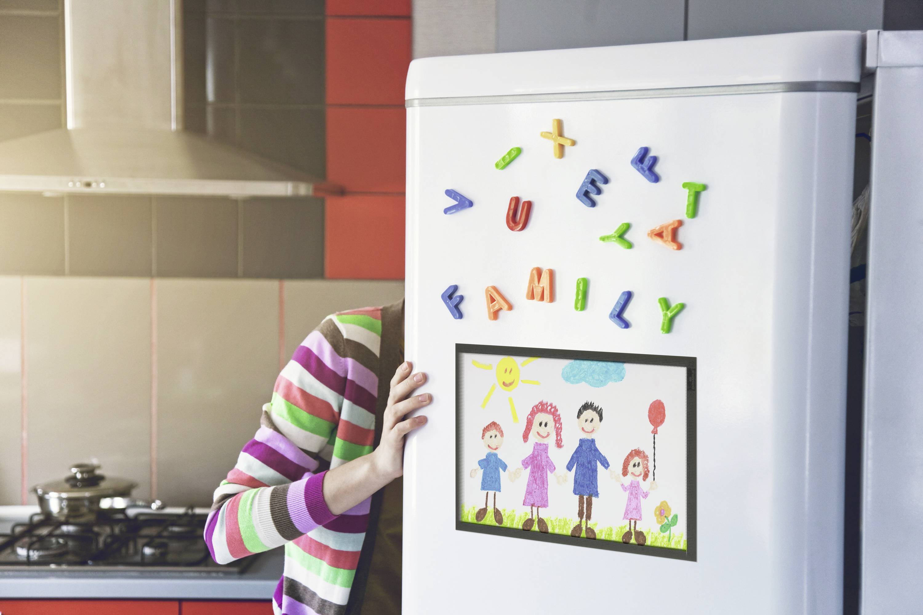 Une personne partiellement cachée derrière un réfrigérateur dans une cuisine, décorée avec des dessins d'enfants et des lettres magnétiques colorées formant le mot 'FAMILY'.
