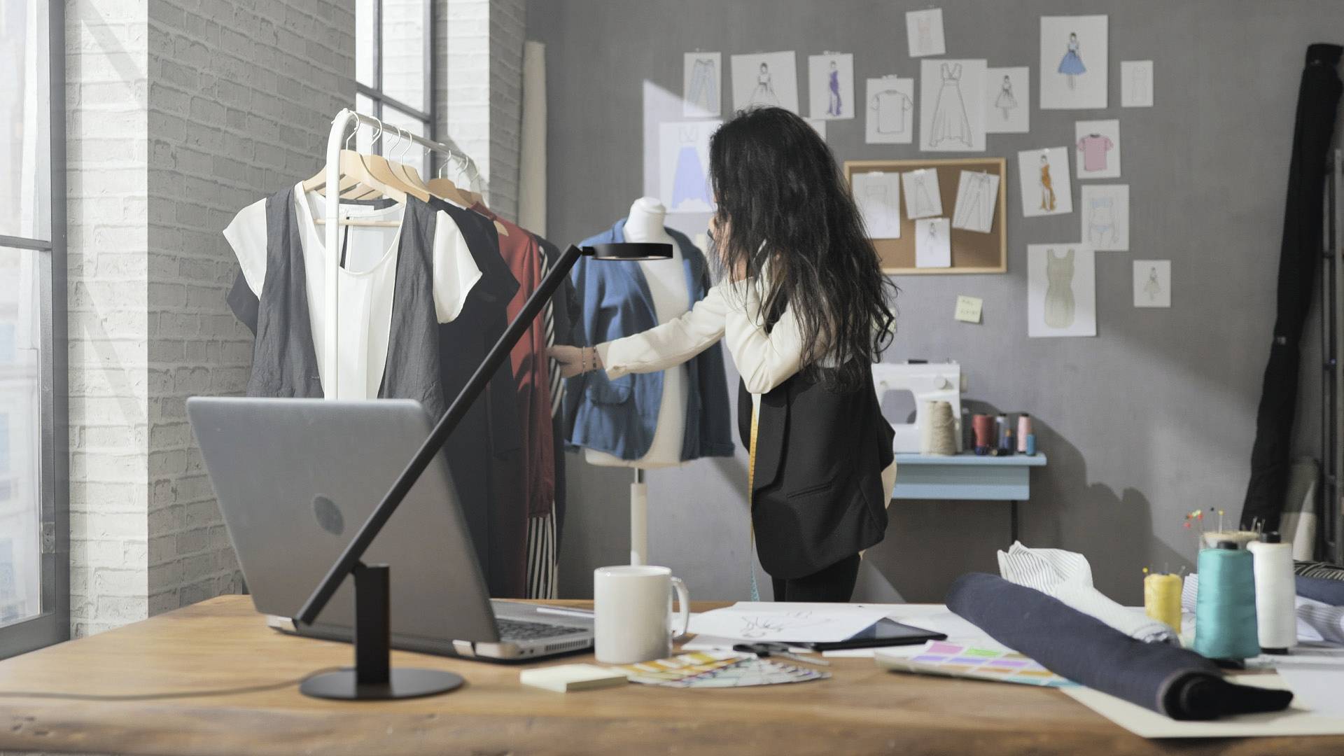 Une designer est au téléphone dans un atelier de mode. Elle examine des vêtements suspendus à un portant. Des croquis sont visibles en arrière-plan.
