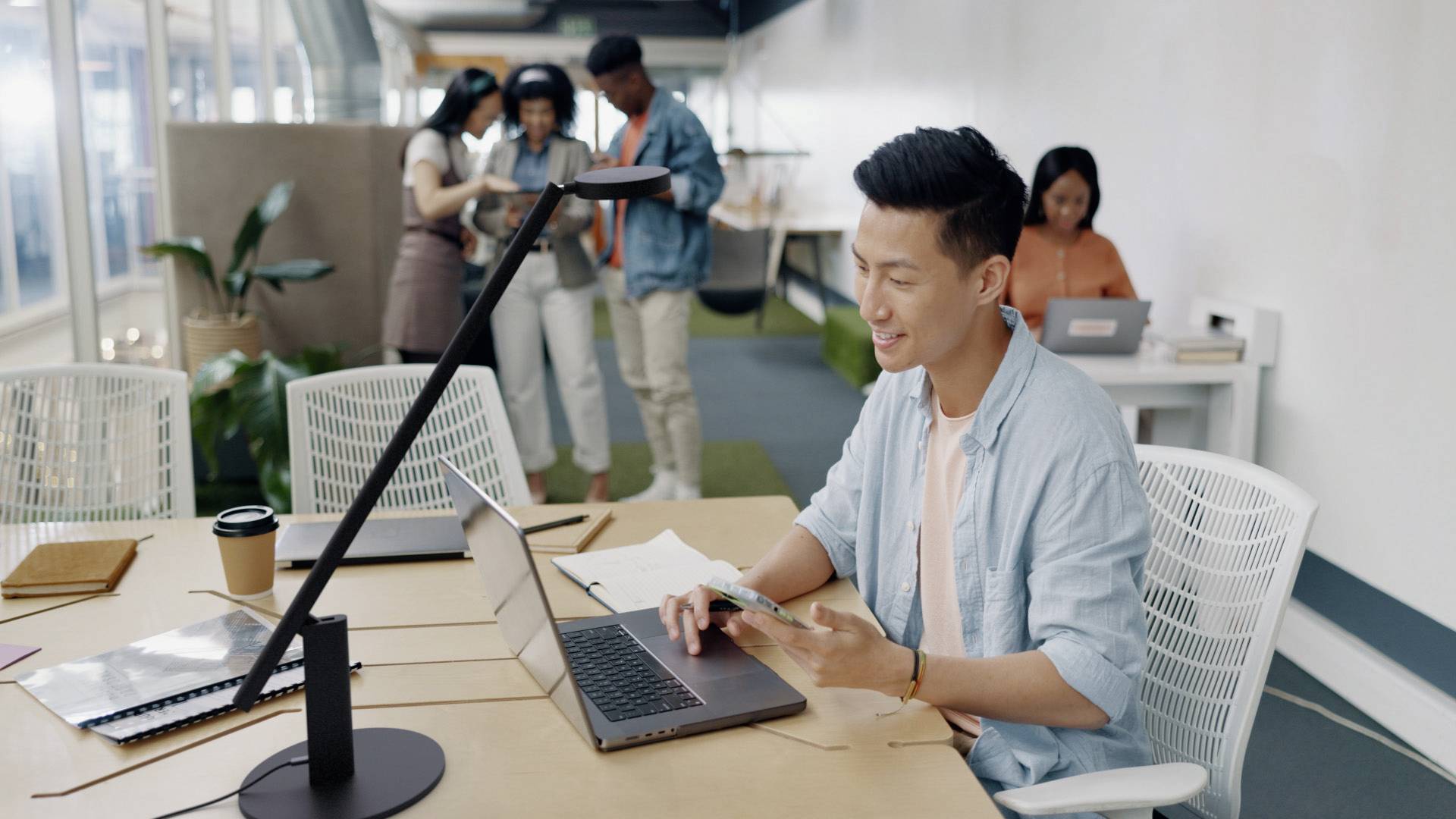 Une personne est assise à un bureau et travaille sur un ordinateur portable. À l'arrière-plan, trois personnes regardent ensemble une tablette.