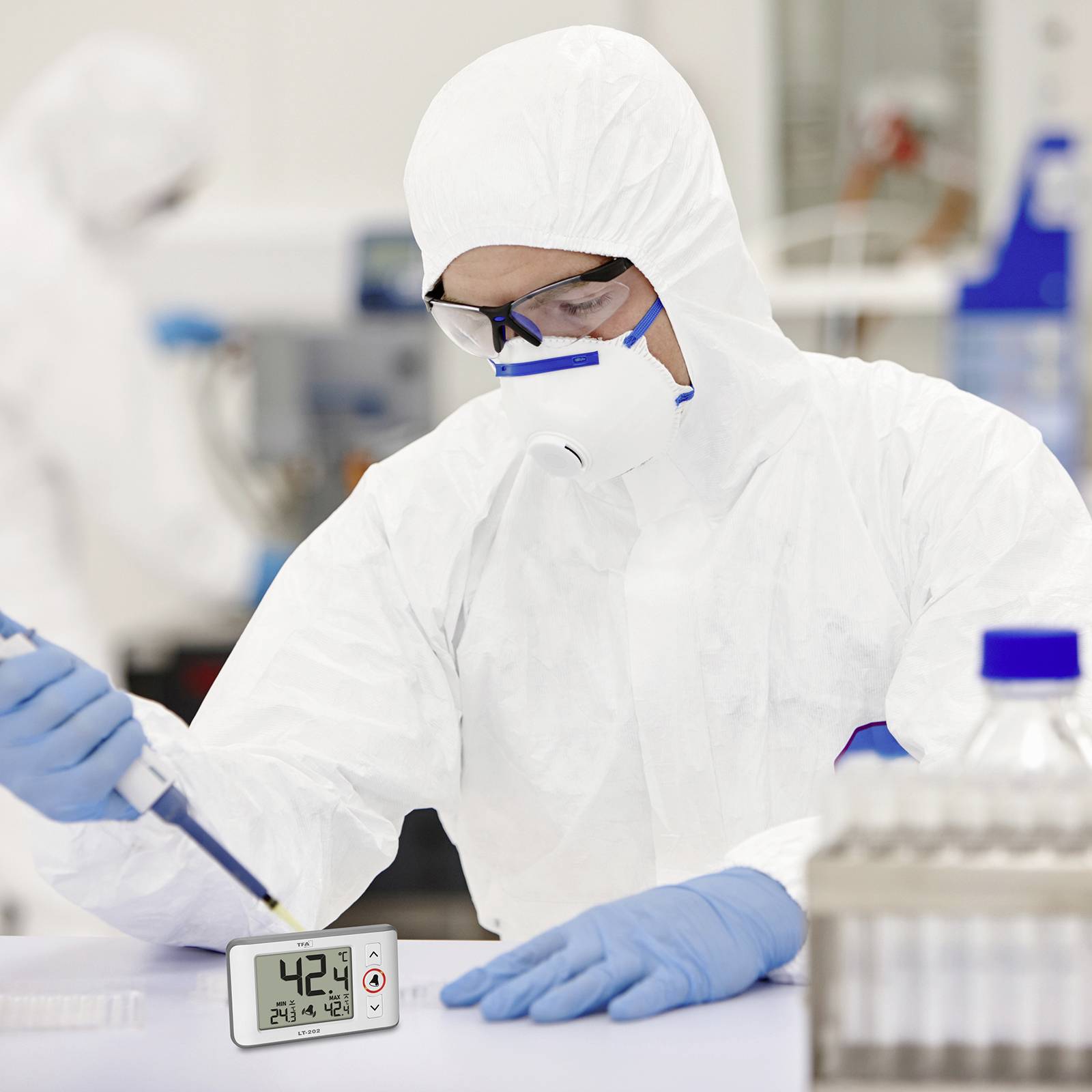 Une personne en combinaison de protection travaille dans un laboratoire, pipette un liquide. Un thermomètre numérique affiche 42,4 °C. Environnement de recherche scientifique.
