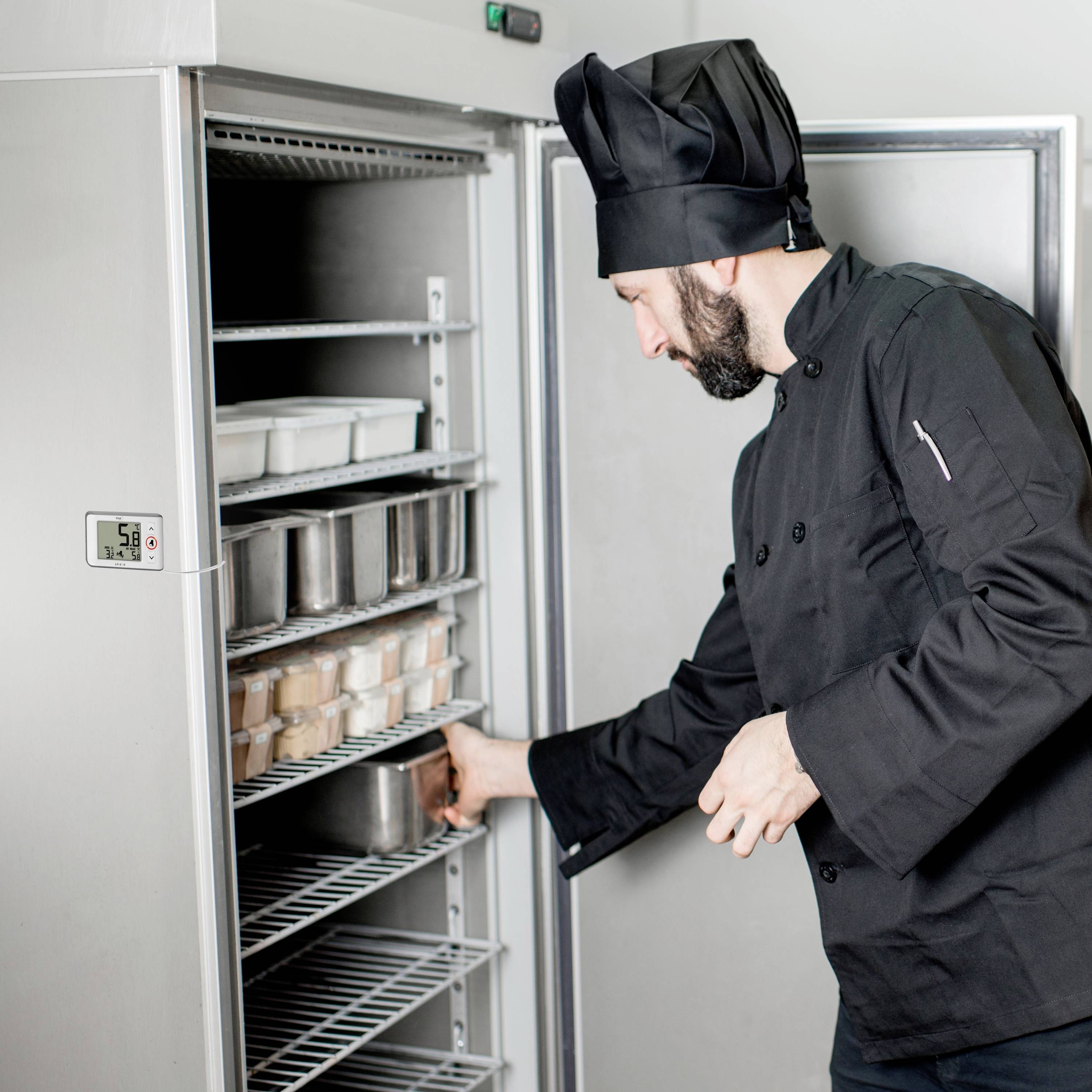 Un cuisinier en uniforme noir range des contenants dans un réfrigérateur en acier inoxydable. Le réfrigérateur est ouvert et rempli de denrées alimentaires.