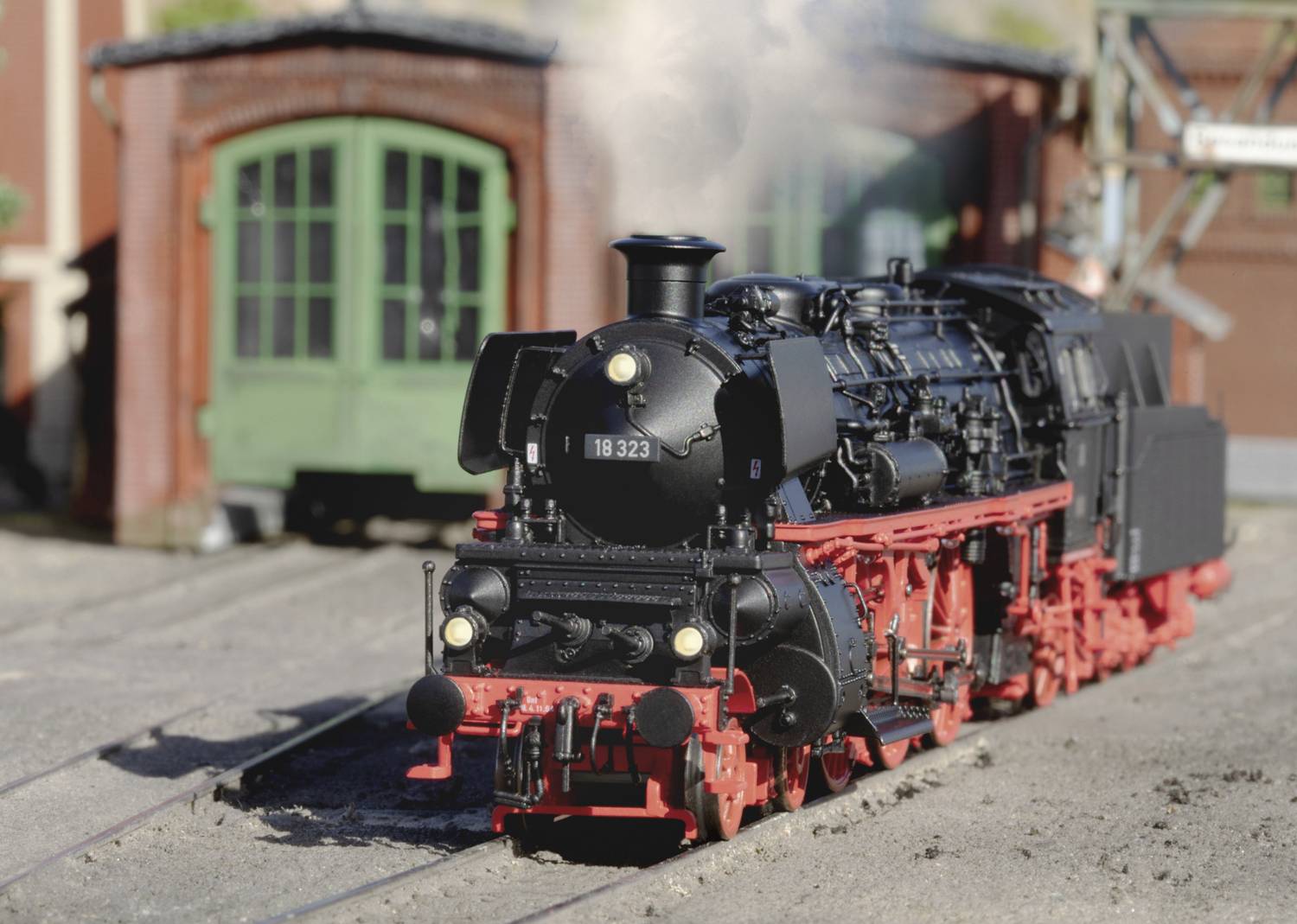 Une locomotive à vapeur noire avec des roues rouges roule sur un rail miniature devant un hangar à locomotives vert en arrière-plan.