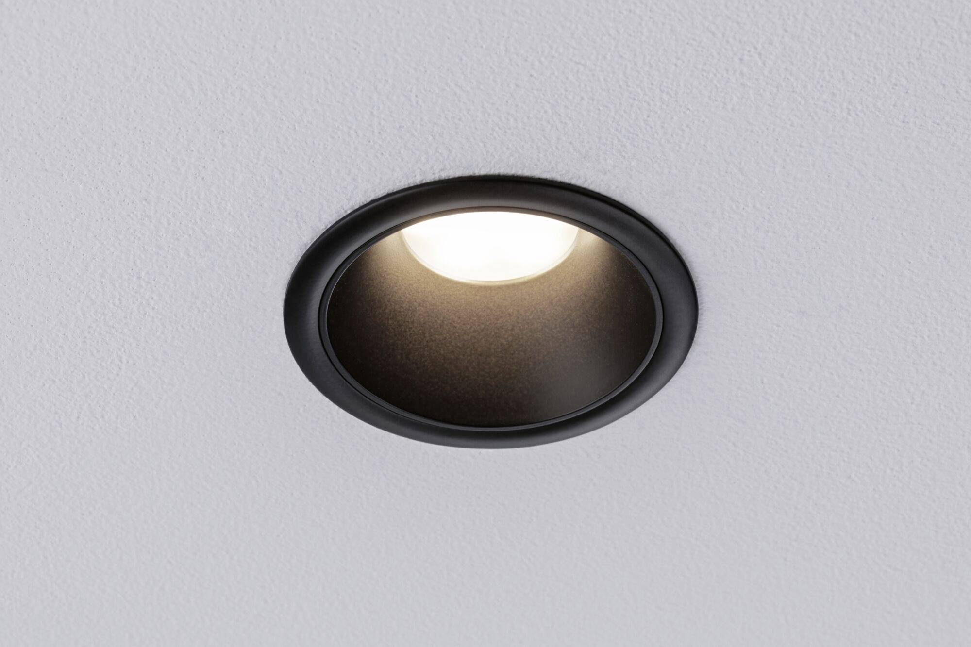 Un luminaire encastré noir et rond dans un plafond blanc, allumé et diffusant une lumière chaleureuse.